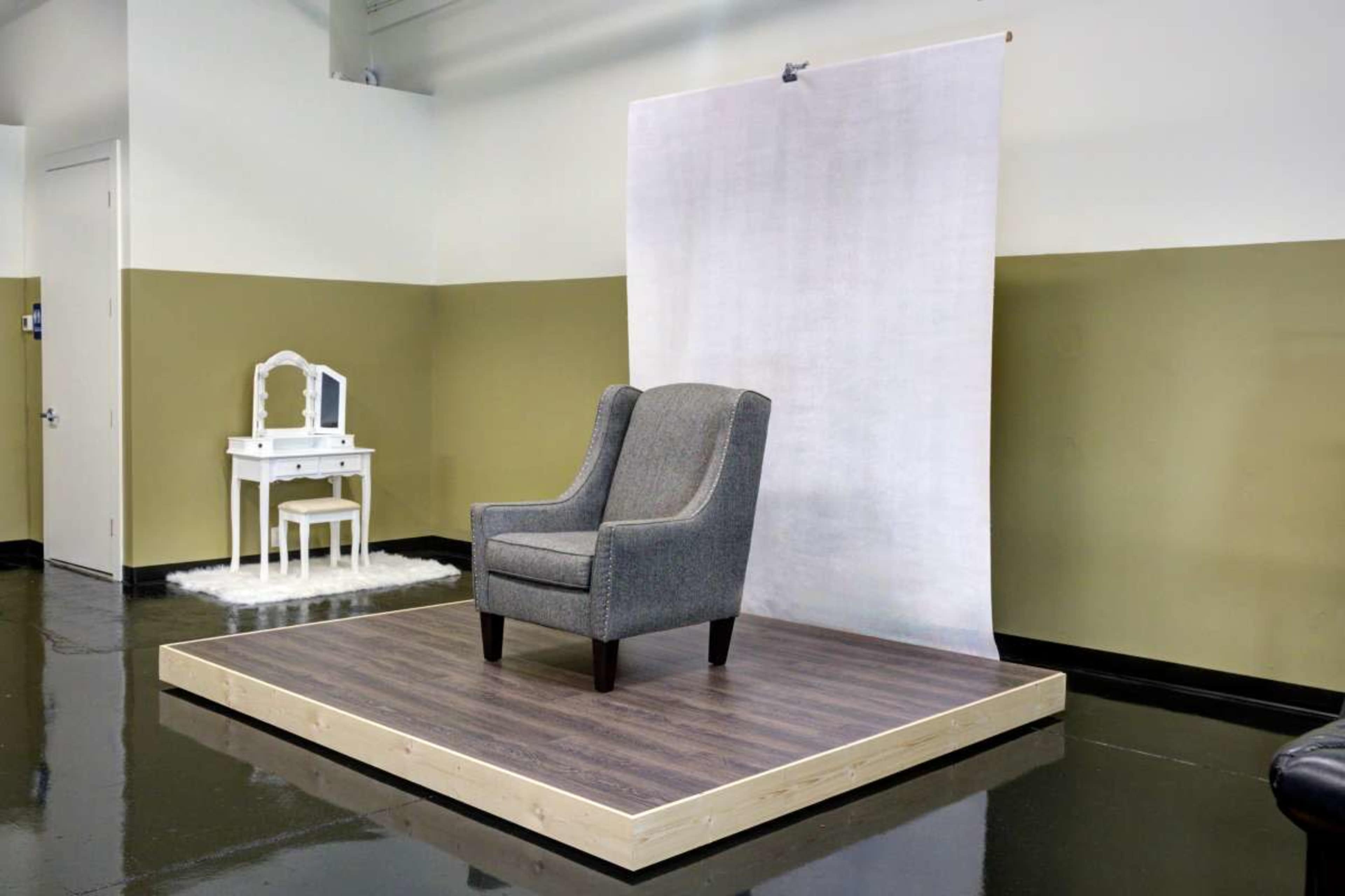 The image shows a gray armchair placed on a wooden platform in a minimalist studio with a white backdrop and a small white vanity table in the background.