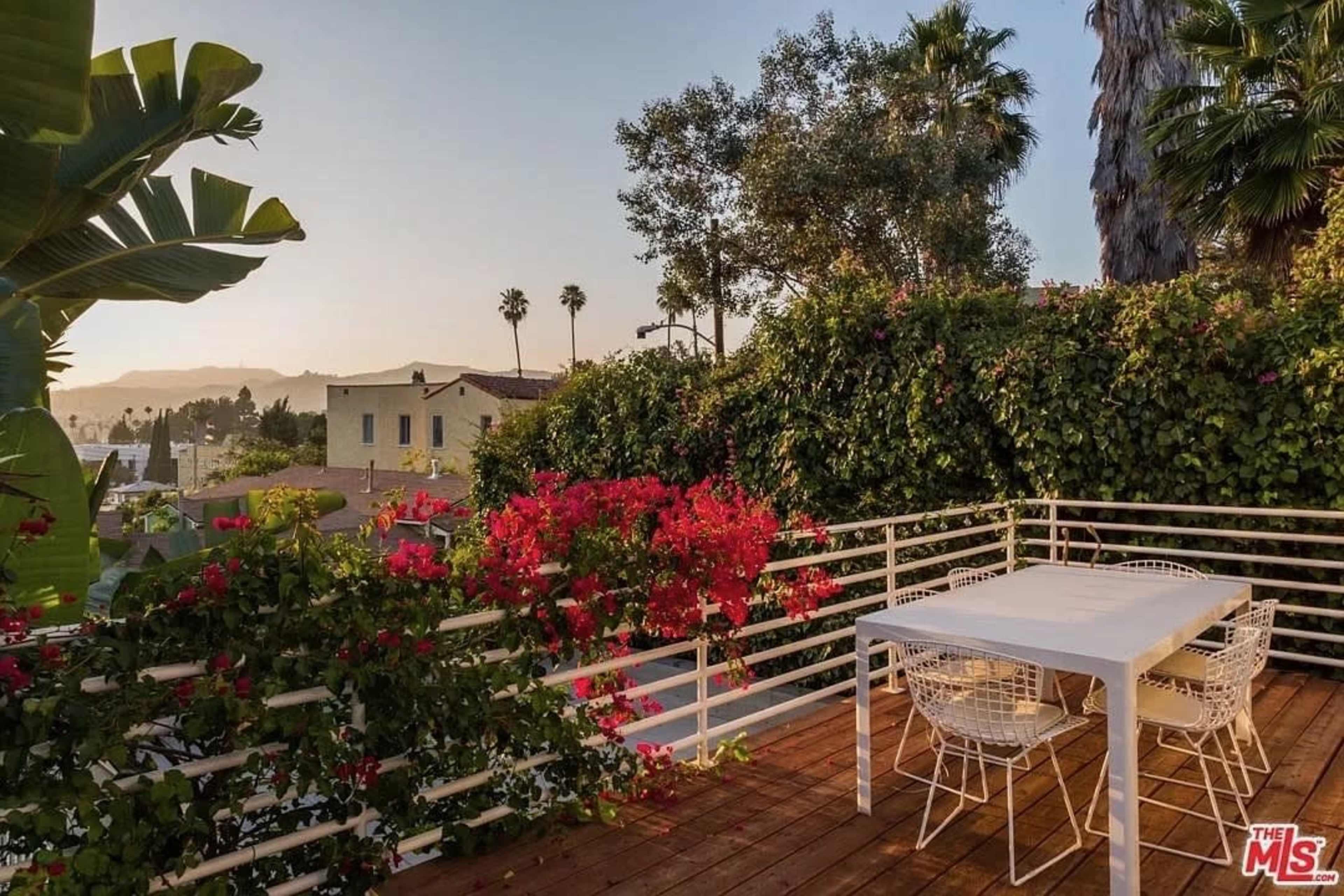 A balcony features a white table and chairs surrounded by vibrant bougainvillea and palm trees, offering a view of the sunset.
