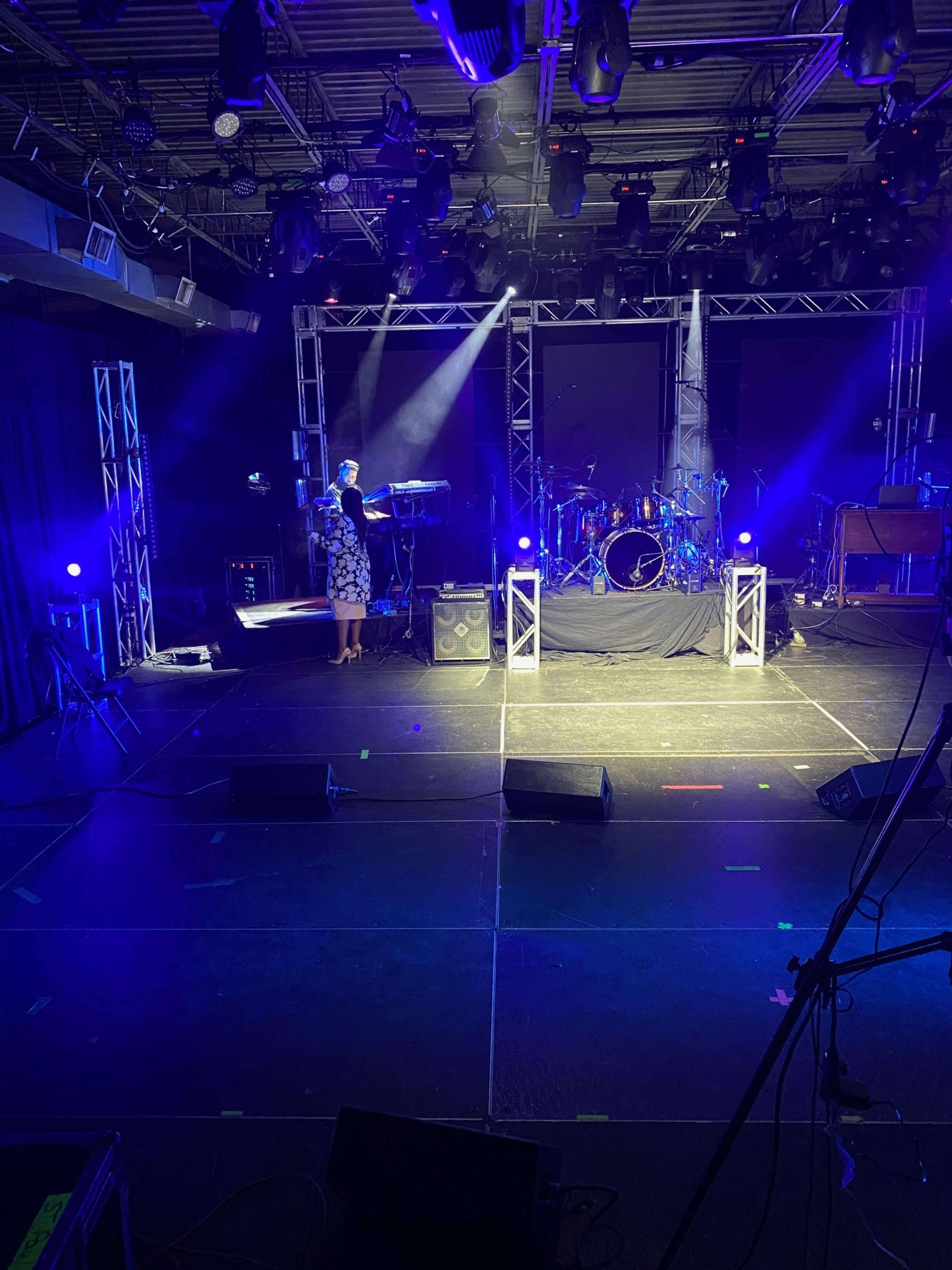 A musician stands on stage in front of a band setup with lights and instruments arranged in a performance space.