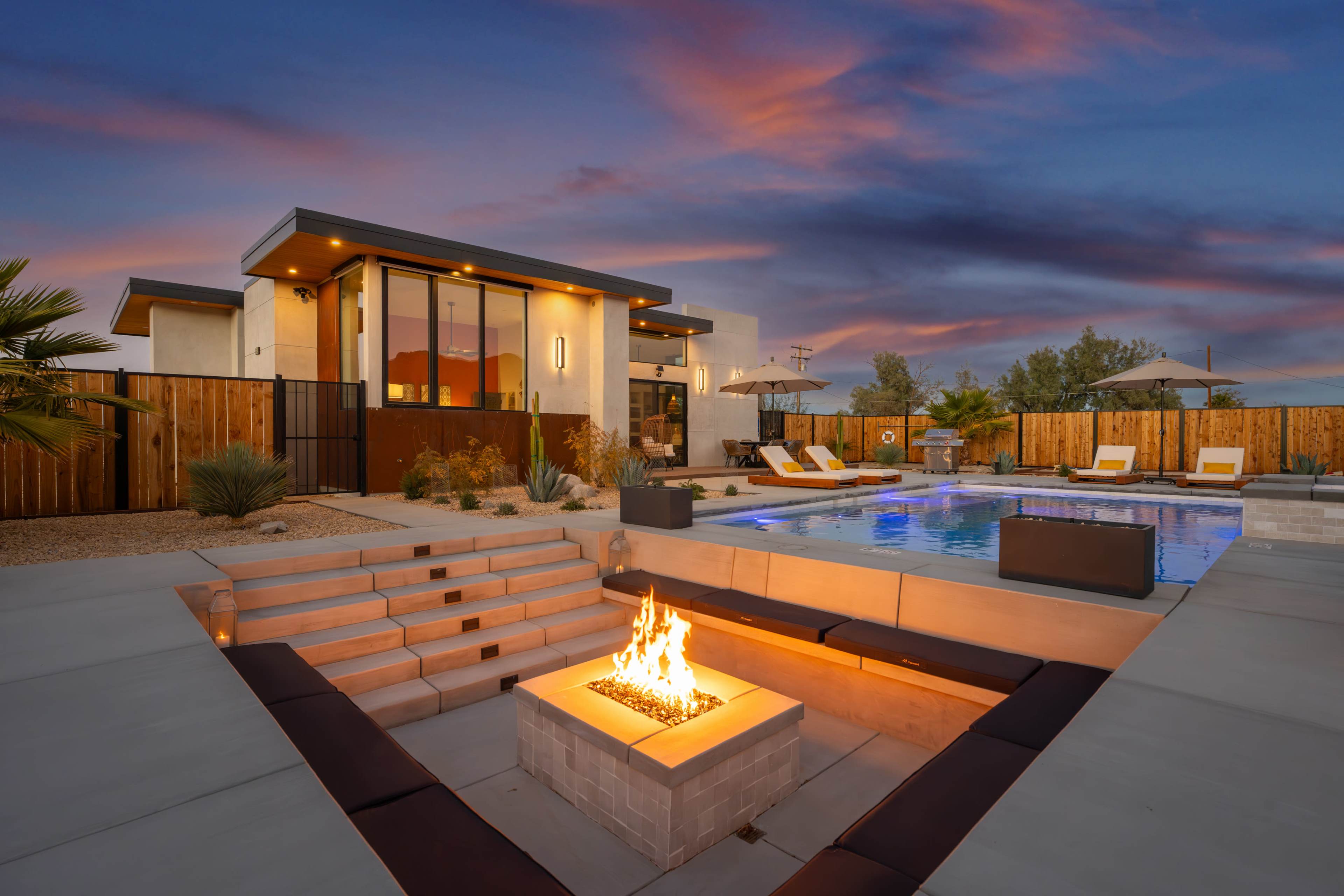 A modern home features a pool and fire pit area, surrounded by landscaping and a wooden fence under a colorful sunset sky.