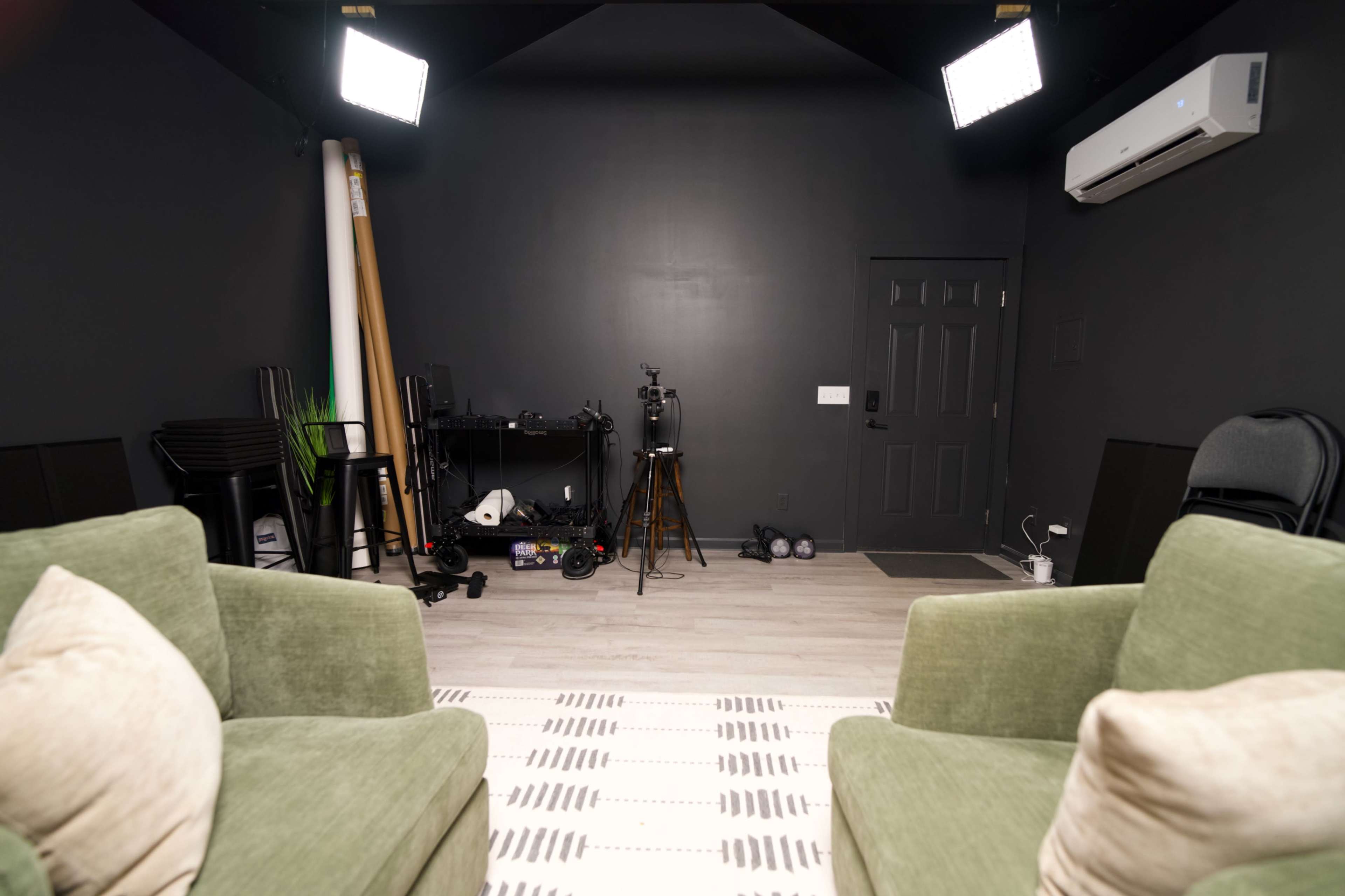 The image shows a dimly lit studio space with dark walls, two green chairs facing each other, and various equipment and furniture arranged in the background.