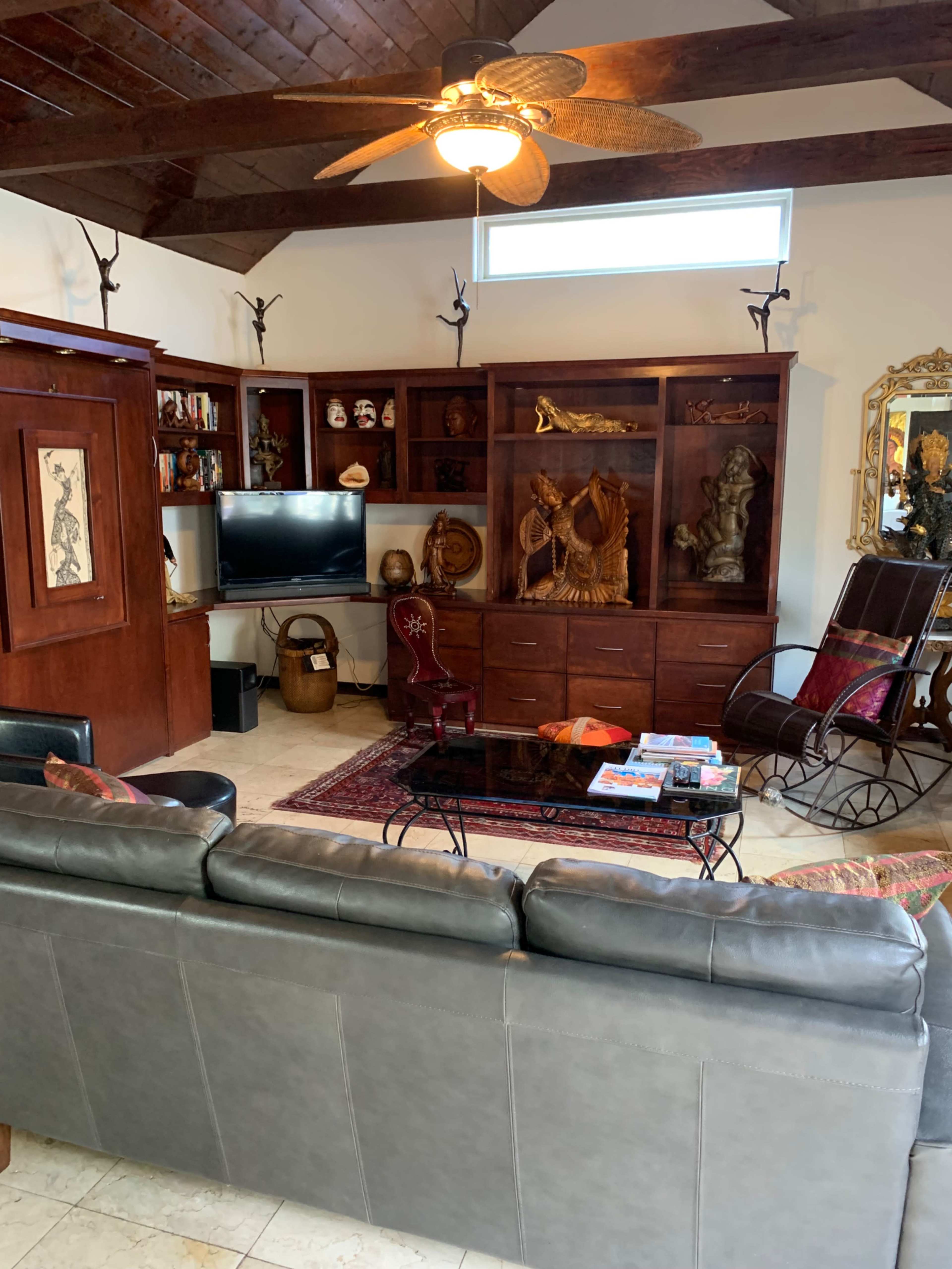 A living room with a leather sofa, a wooden cabinet filled with decorative items, a television, and a rocking chair, all under a ceiling fan.