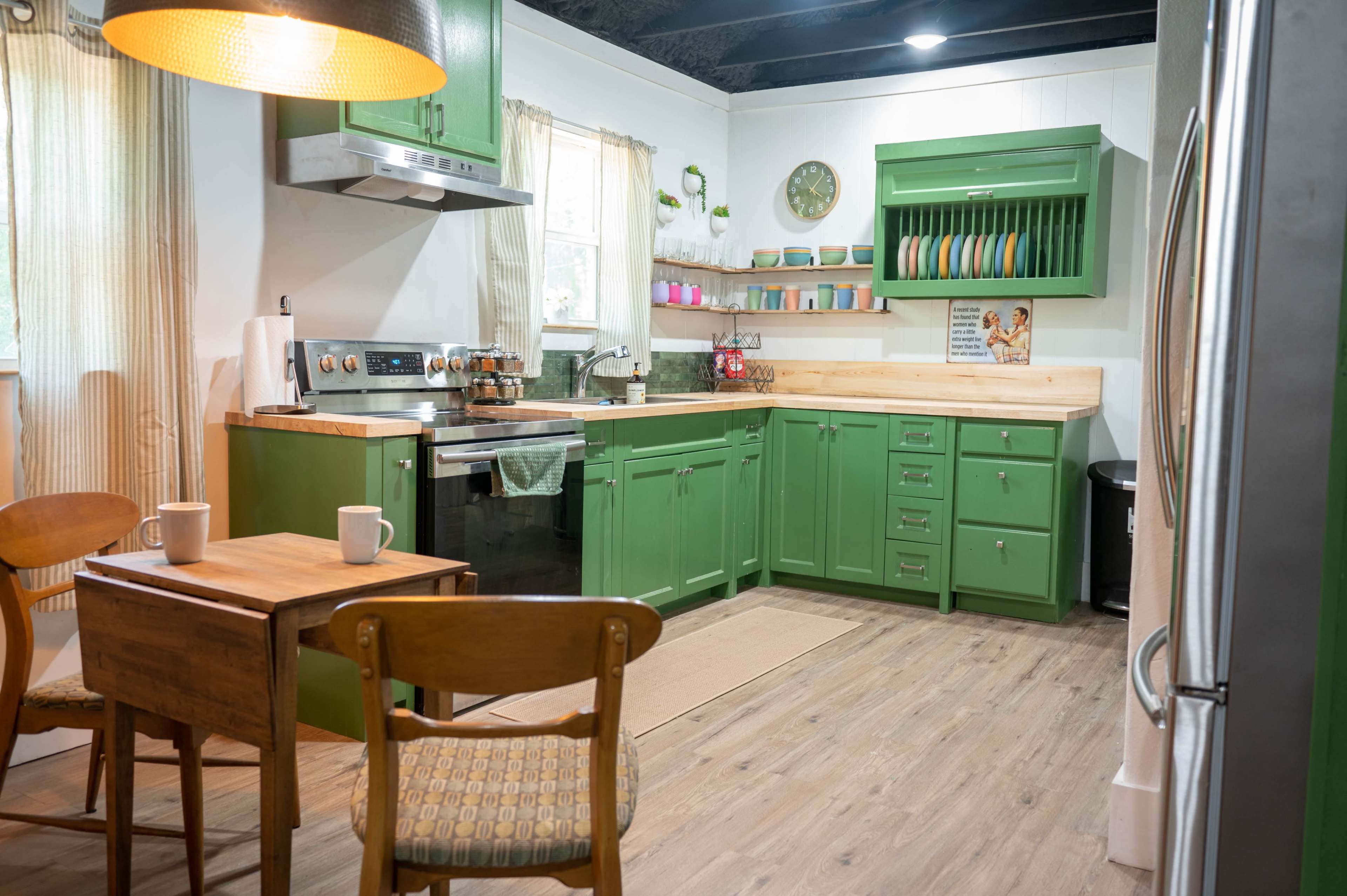 The image shows a kitchen with green cabinetry, a wooden dining table, and two chairs, featuring a stove, refrigerator, and an arrangement of colorful dishware on open shelves.