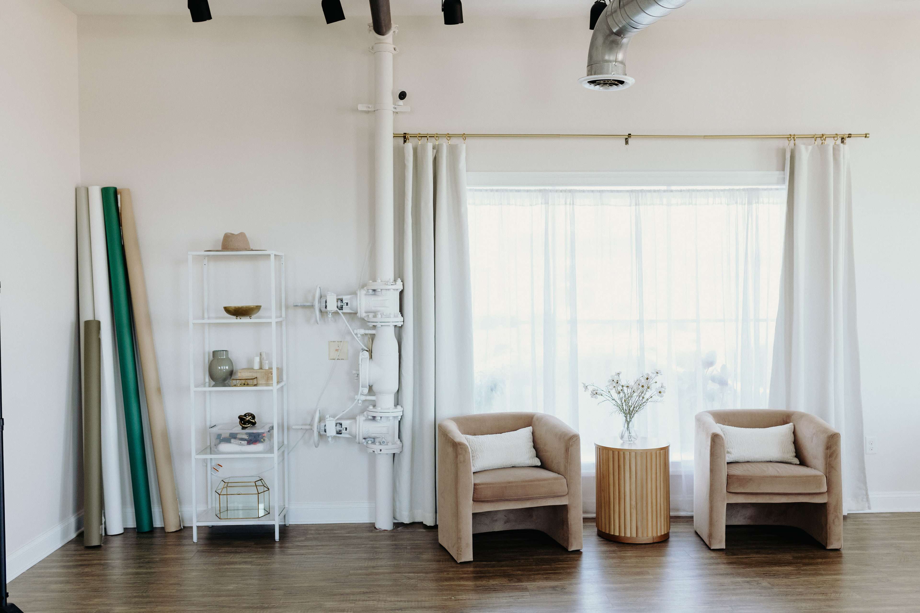 A bright room features two beige armchairs next to a round wooden table, with a clear shelving unit displaying various items and a window covered by sheer white curtains.