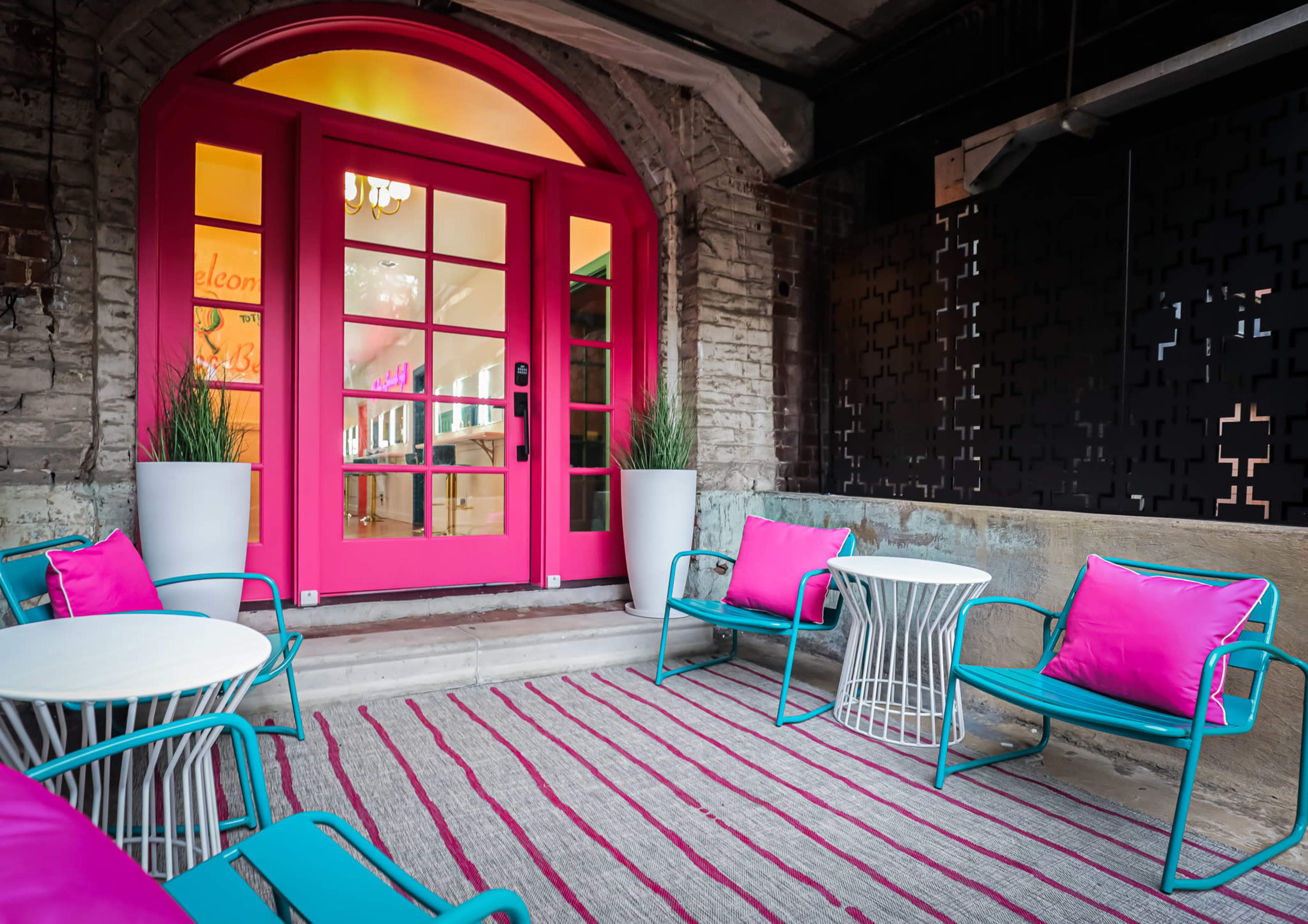 A brightly colored entrance with a pink door surrounded by outdoor seating that includes blue chairs and white tables on a striped rug.