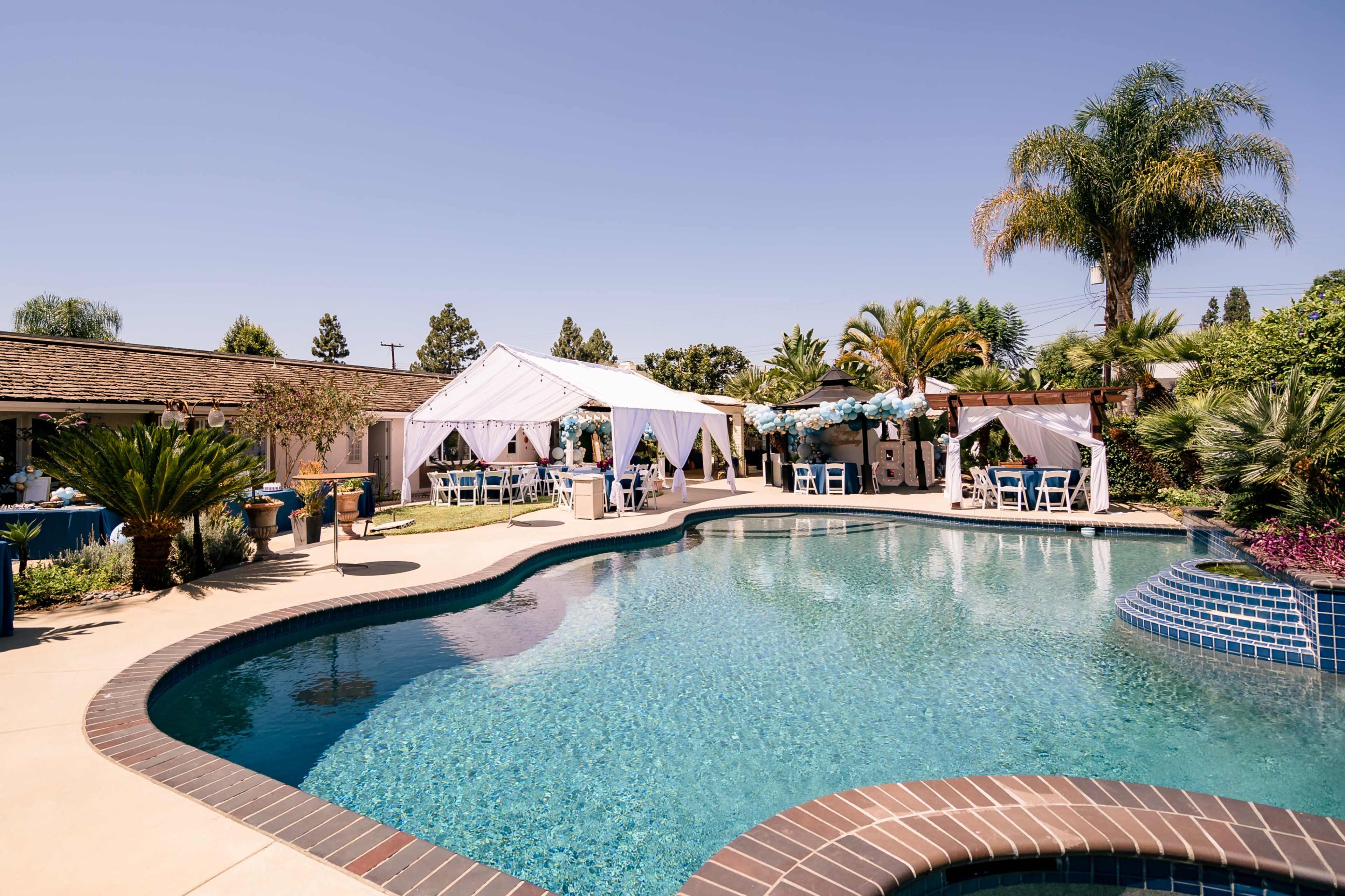 A clear swimming pool is surrounded by tents and seating in a landscaped backyard.