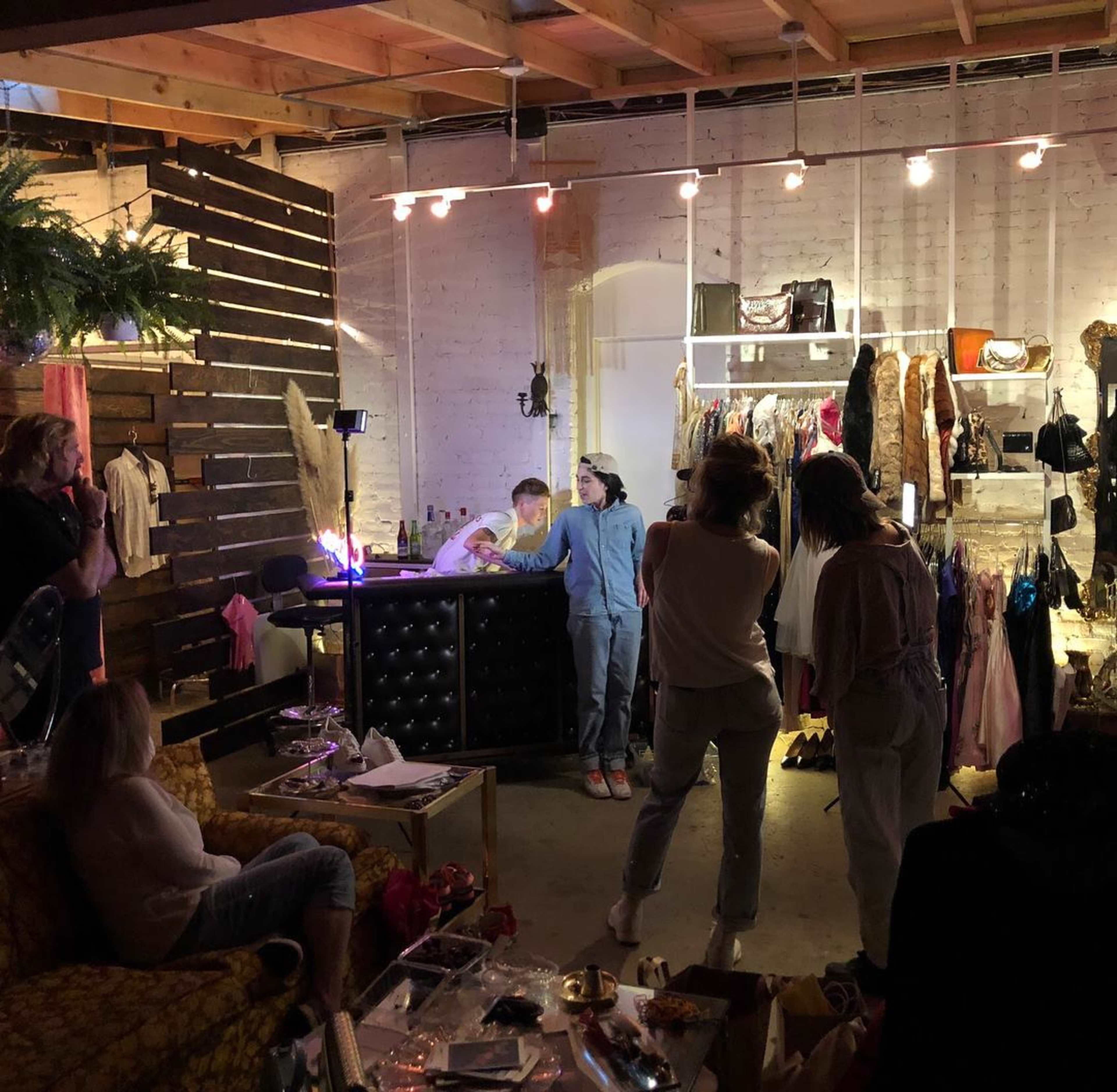 A group of people is gathered in a boutique setting, where clothing racks and a stylish counter are visible amid various fashion items and decor.