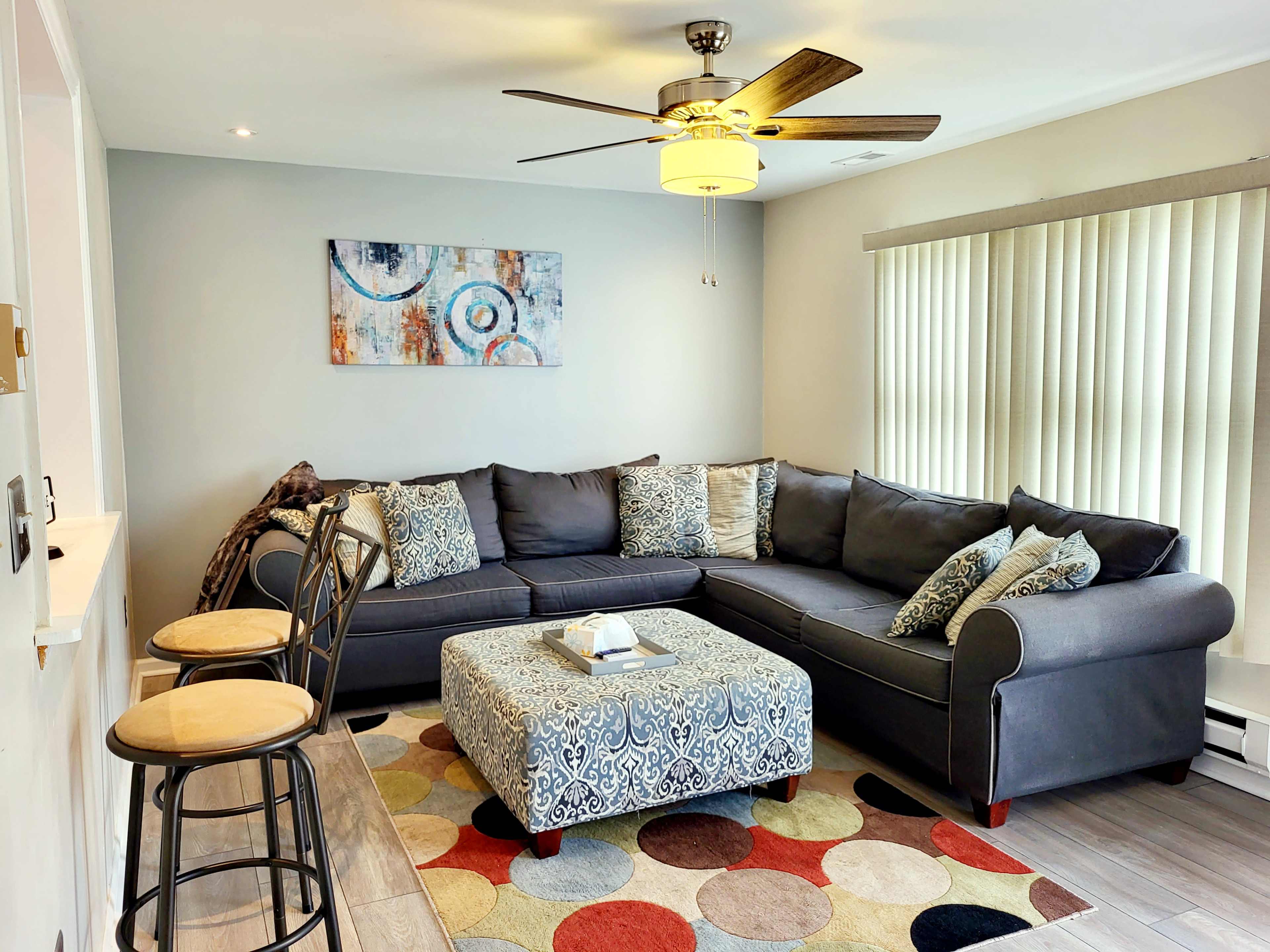 The living room features a gray sectional sofa with decorative pillows, an ottoman, a circular rug, and a ceiling fan, arranged around a bright window with vertical blinds.