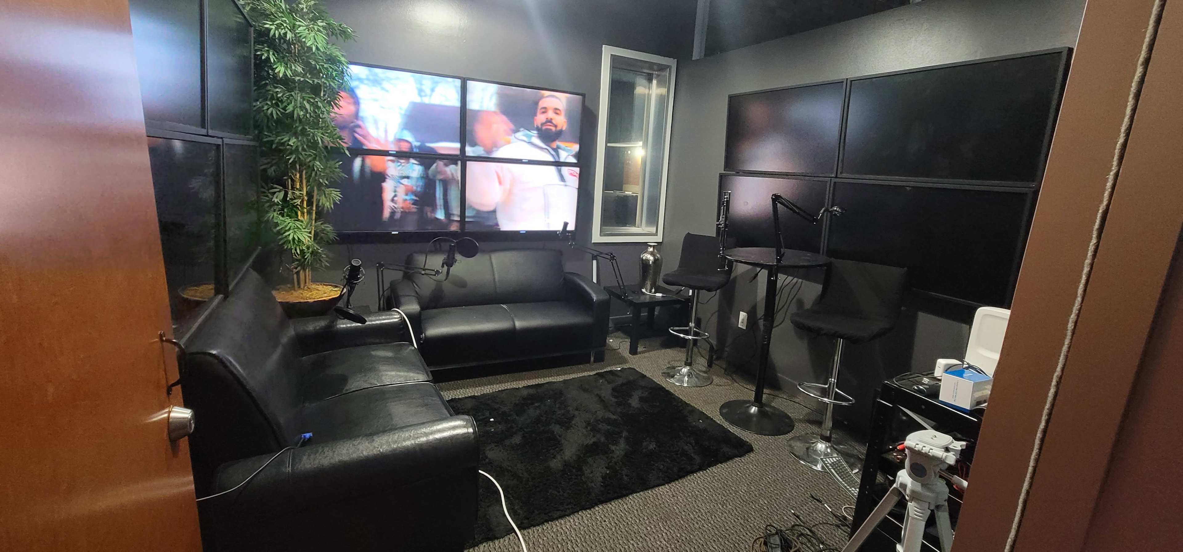 A small, modern recording space featuring a black couch, bar stools, and a large screen displaying video content.