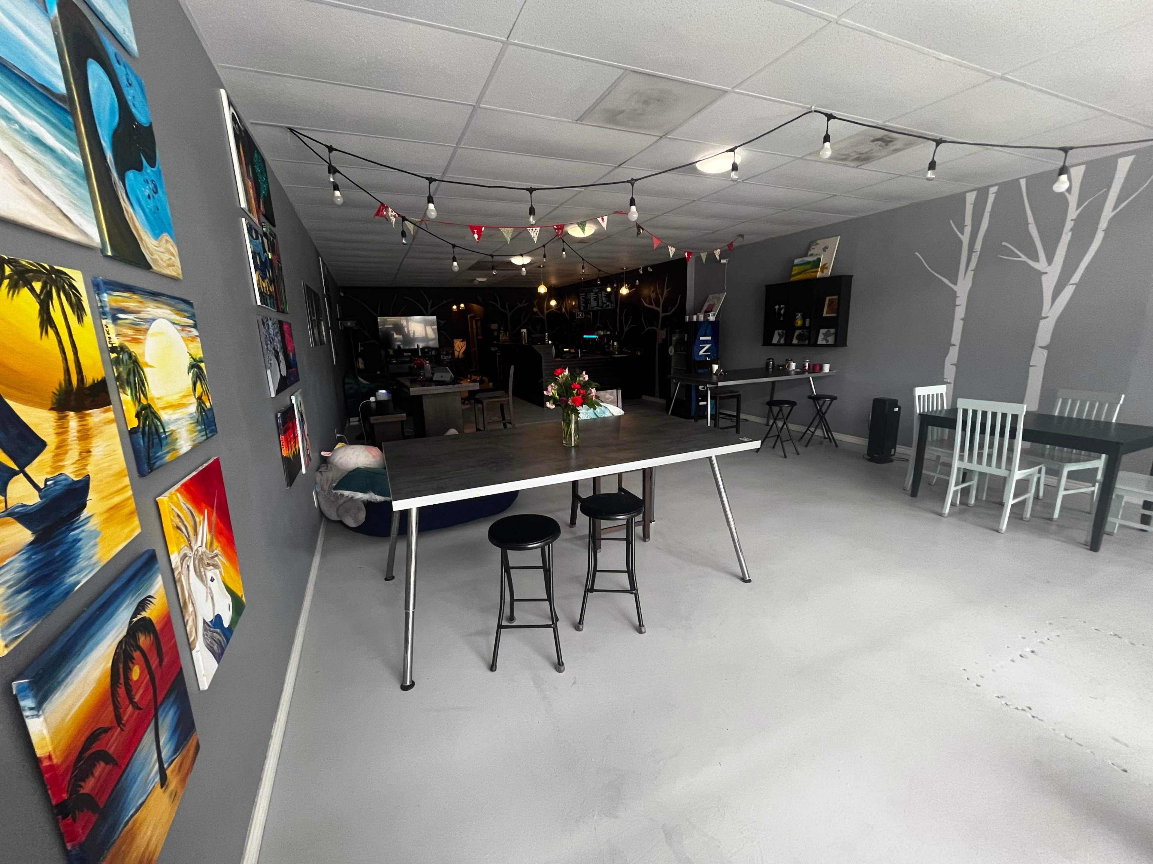 A spacious, modern interior featuring a large table with two stools, decorative wall art, and string lights overhead.