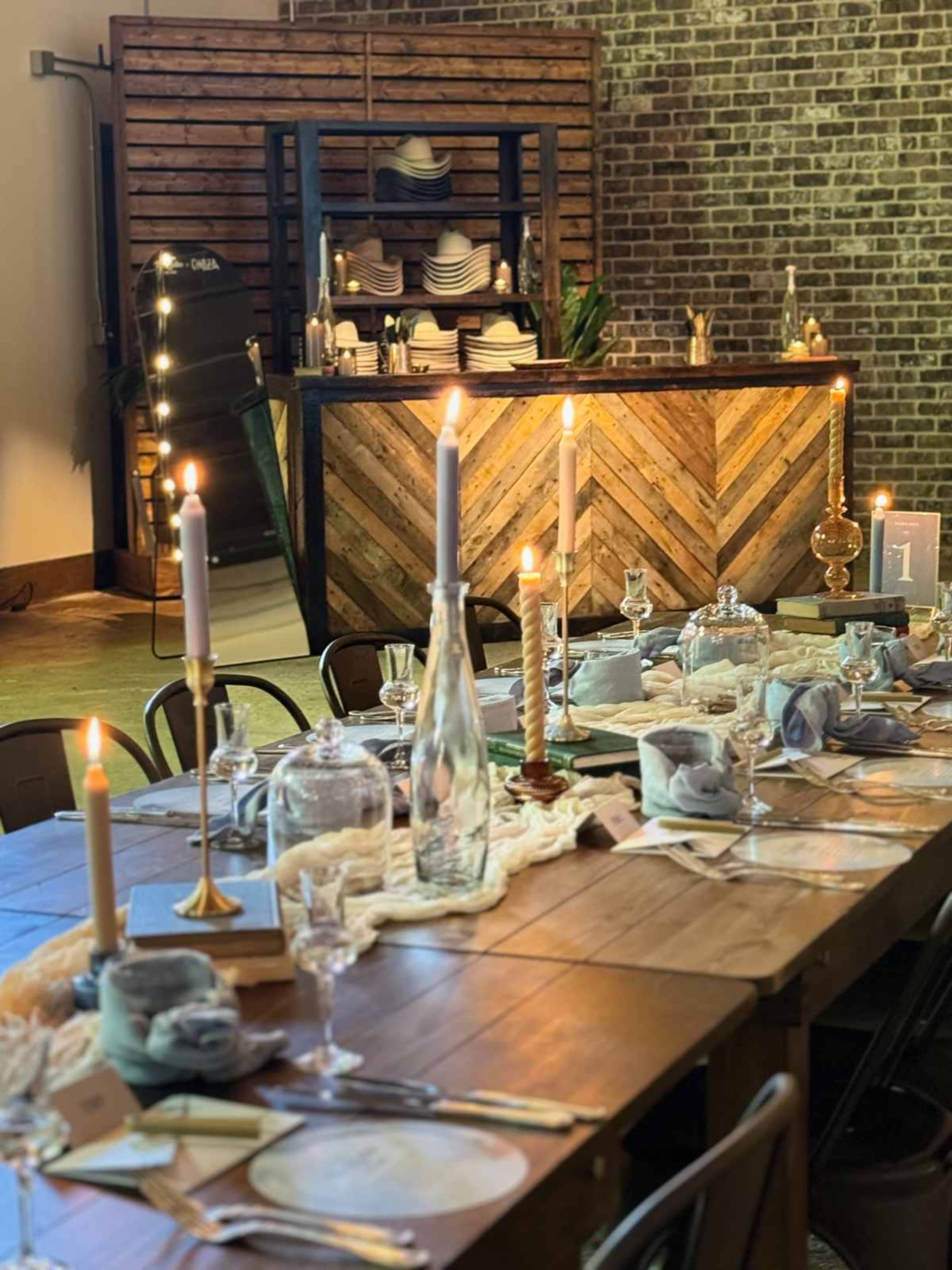 The image shows a dining table set with elegant tableware and candles, complemented by a rustic wooden bar and a brick wall in the background.