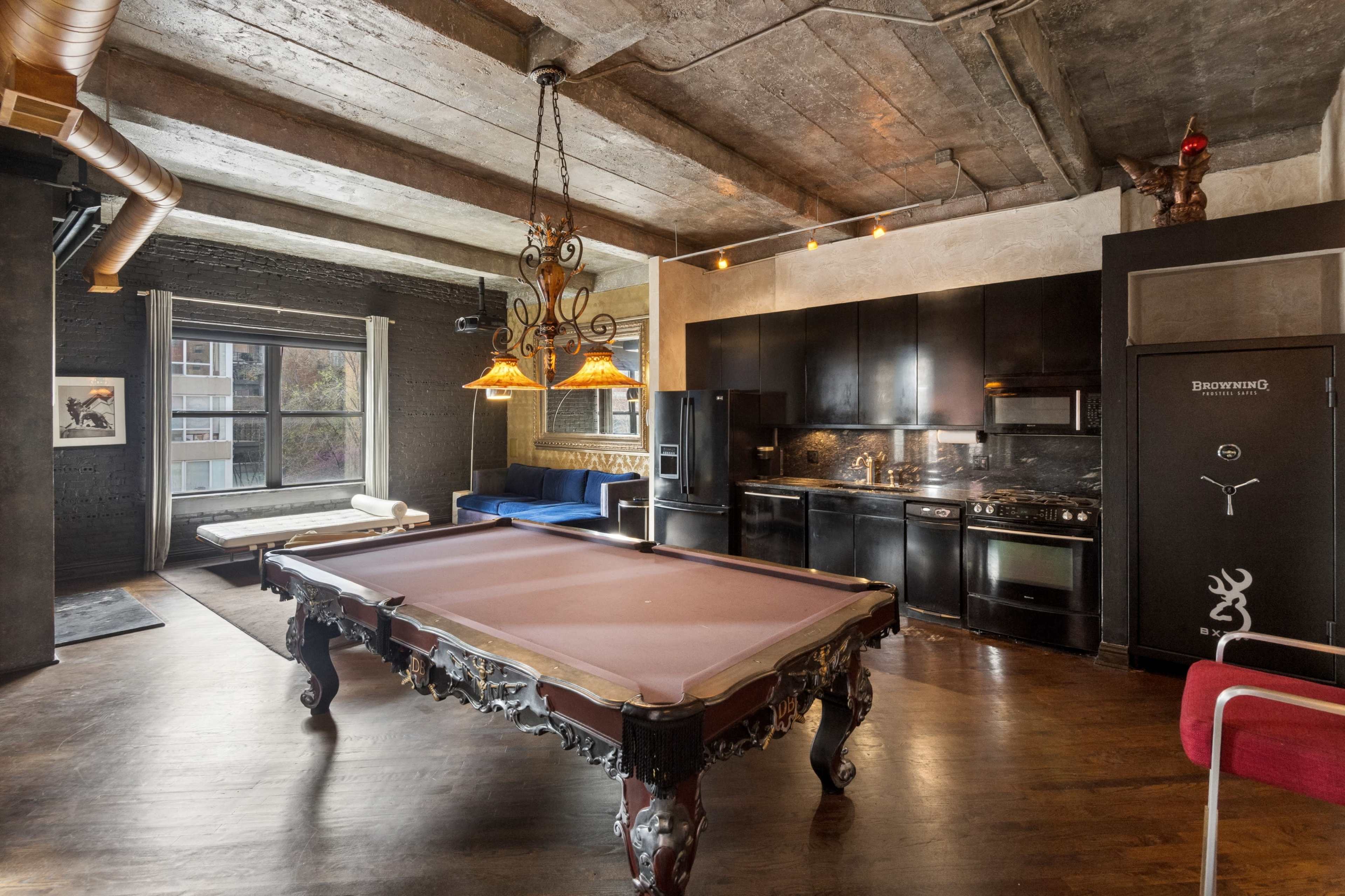 The image shows an industrial-style room featuring a pool table, a kitchenette with black appliances, and a large window allowing natural light to enter.