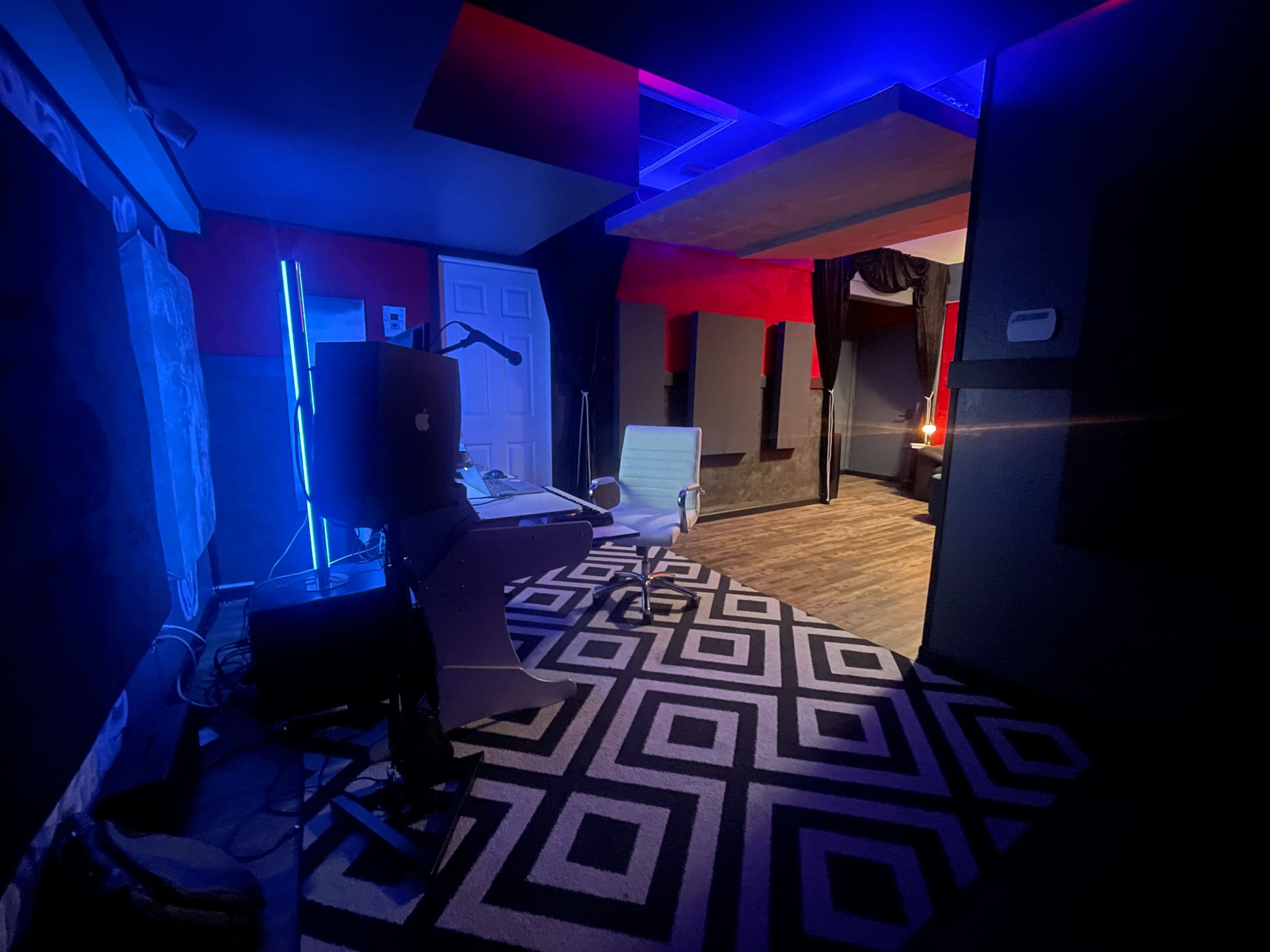 The image shows a dimly lit modern workspace featuring a desk with a computer, a chair, and a geometric patterned rug.