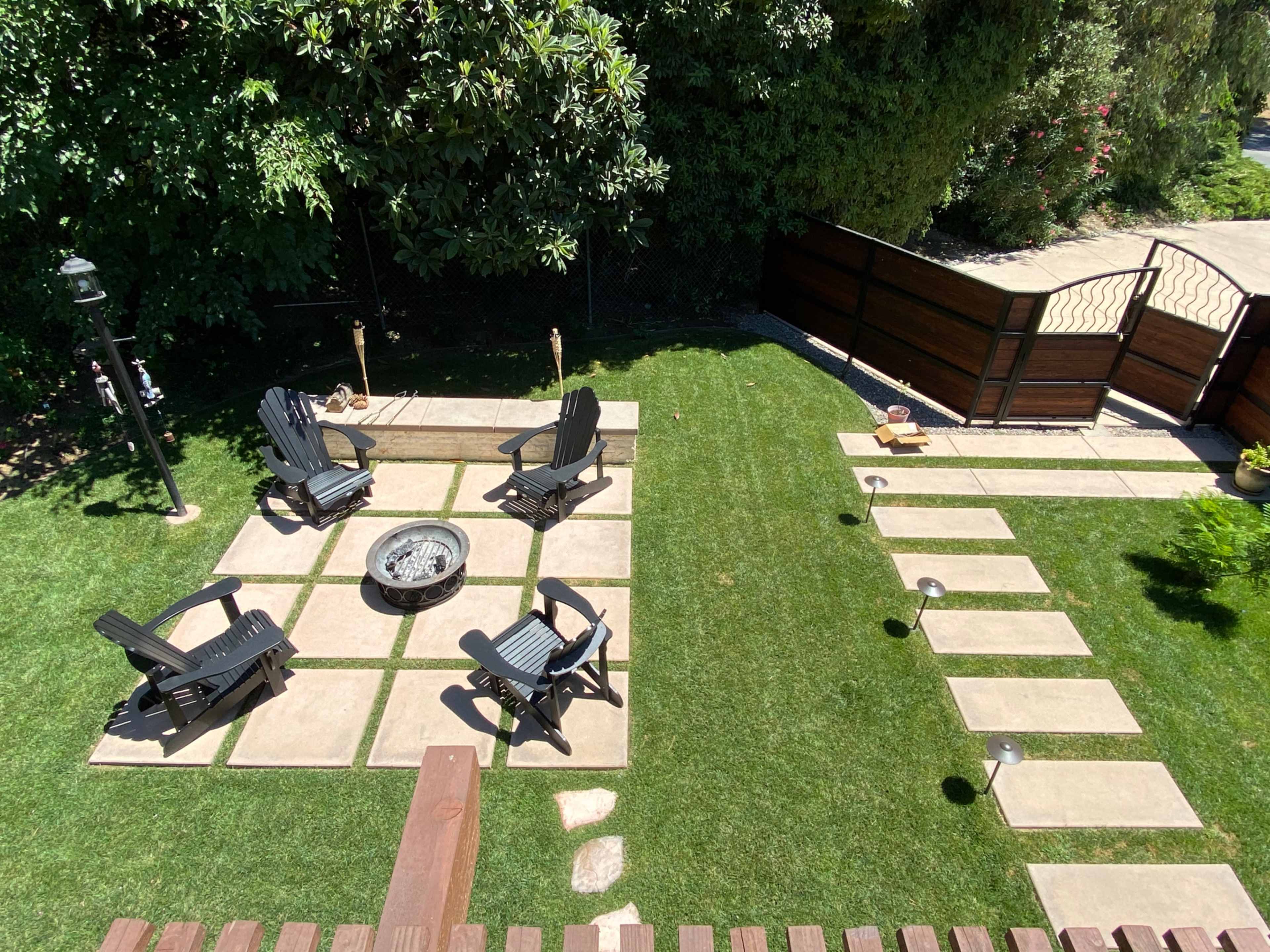 A landscaped backyard featuring a circular stone fire pit surrounded by four black Adirondack chairs, with a pathway of stone pavers leading through a grassy area.