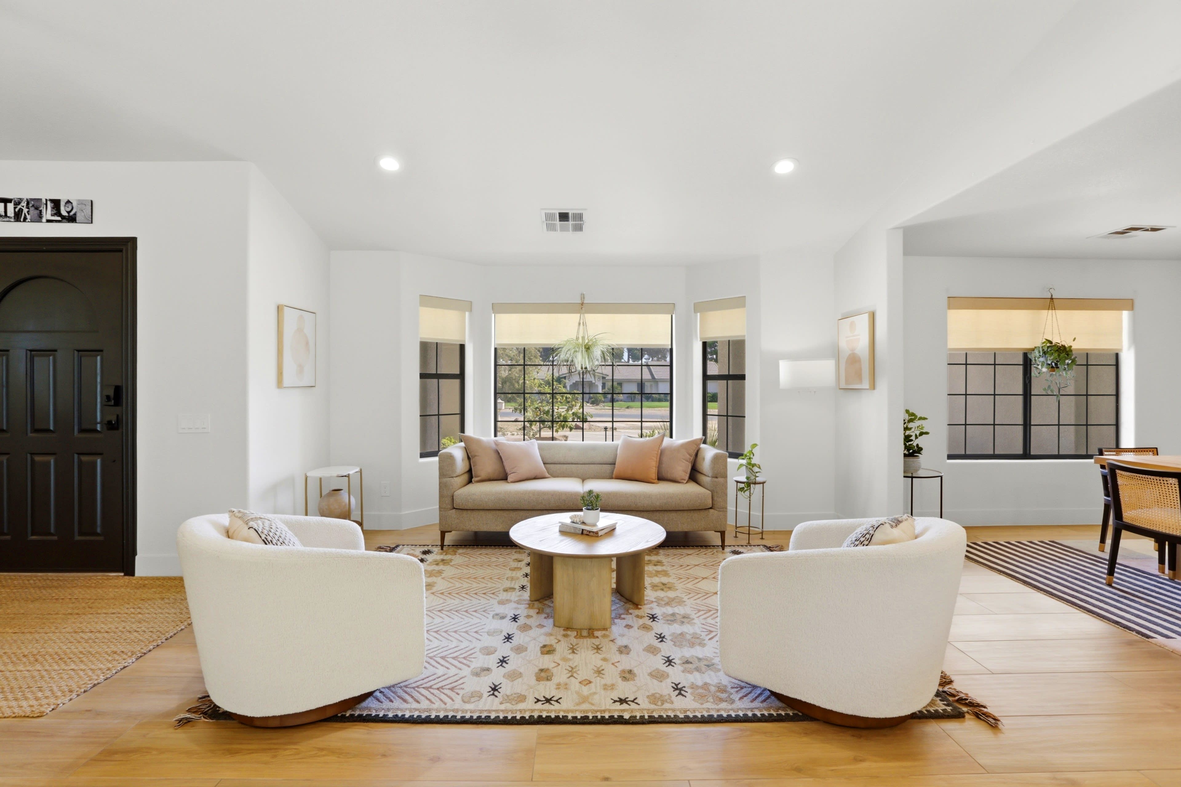 A modern living room features a beige sofa and two white armchairs arranged around a round coffee table, with large windows providing natural light.