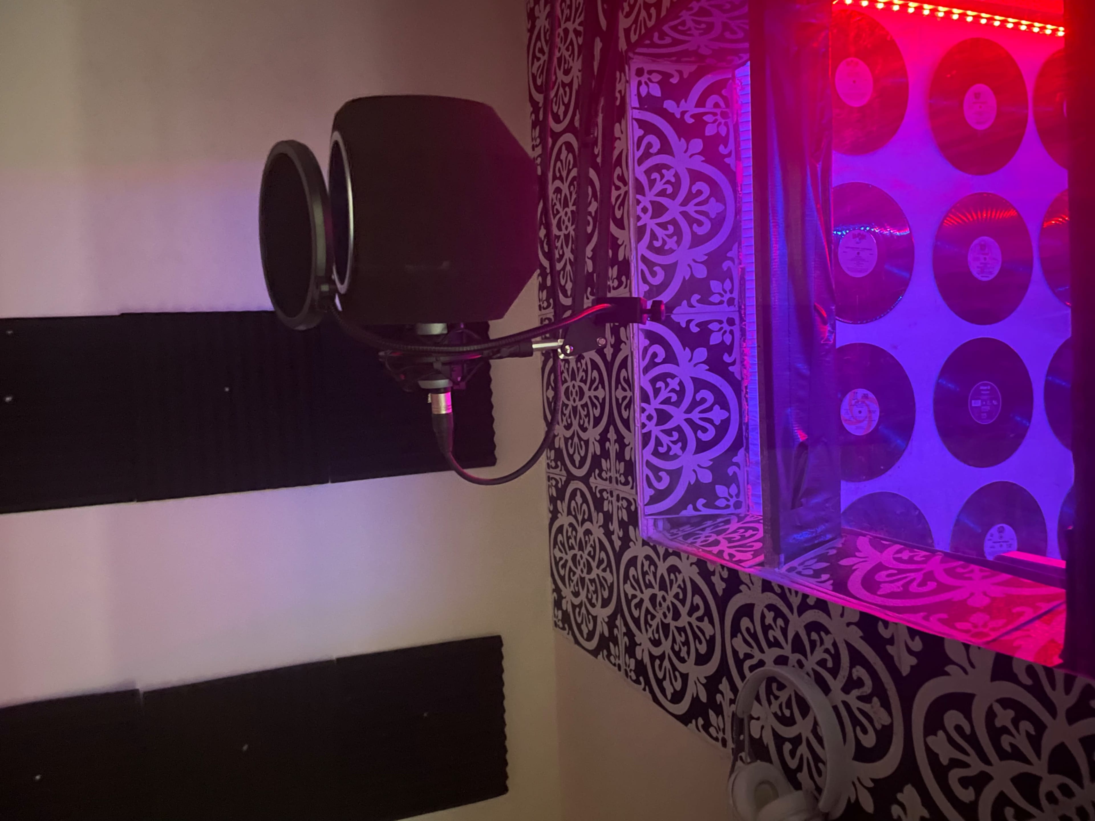 A recording studio featuring a microphone mounted on an arm, soundproofing panels on the walls, and a window displaying circular objects illuminated by red and blue lighting.