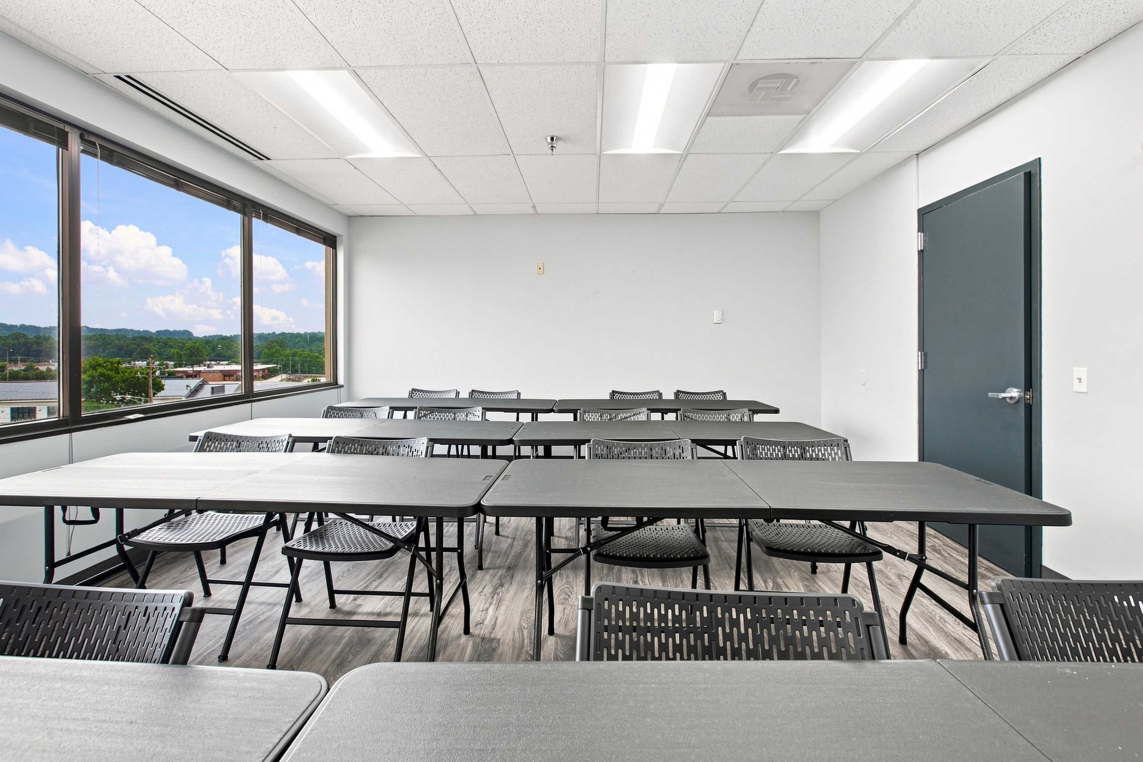 The image shows a clean, well-lit classroom or meeting space with several tables and chairs arranged neatly, under bright overhead lights and with large windows providing a view of the outdoors.