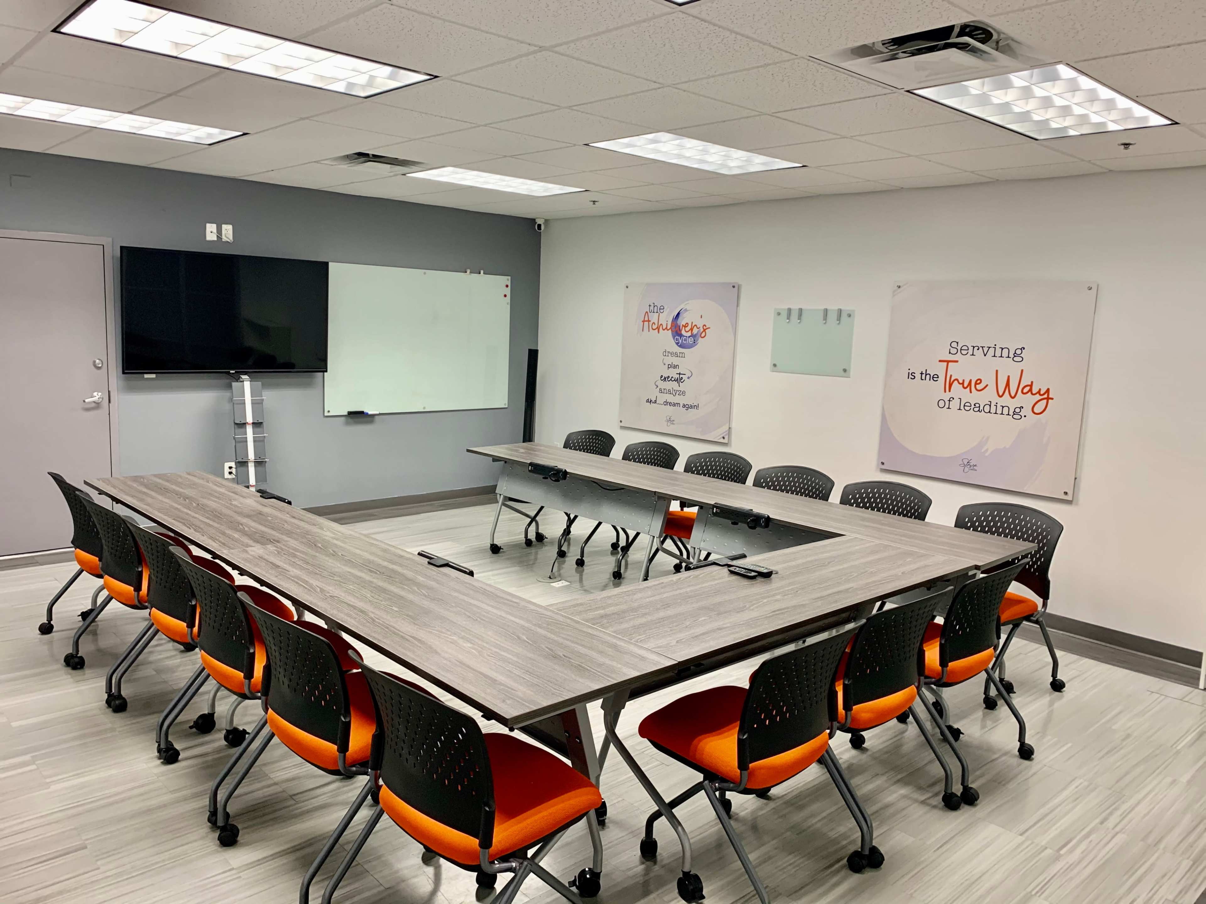 A meeting room features a large triangular table surrounded by black and orange chairs, with whiteboards and wall art displayed nearby.