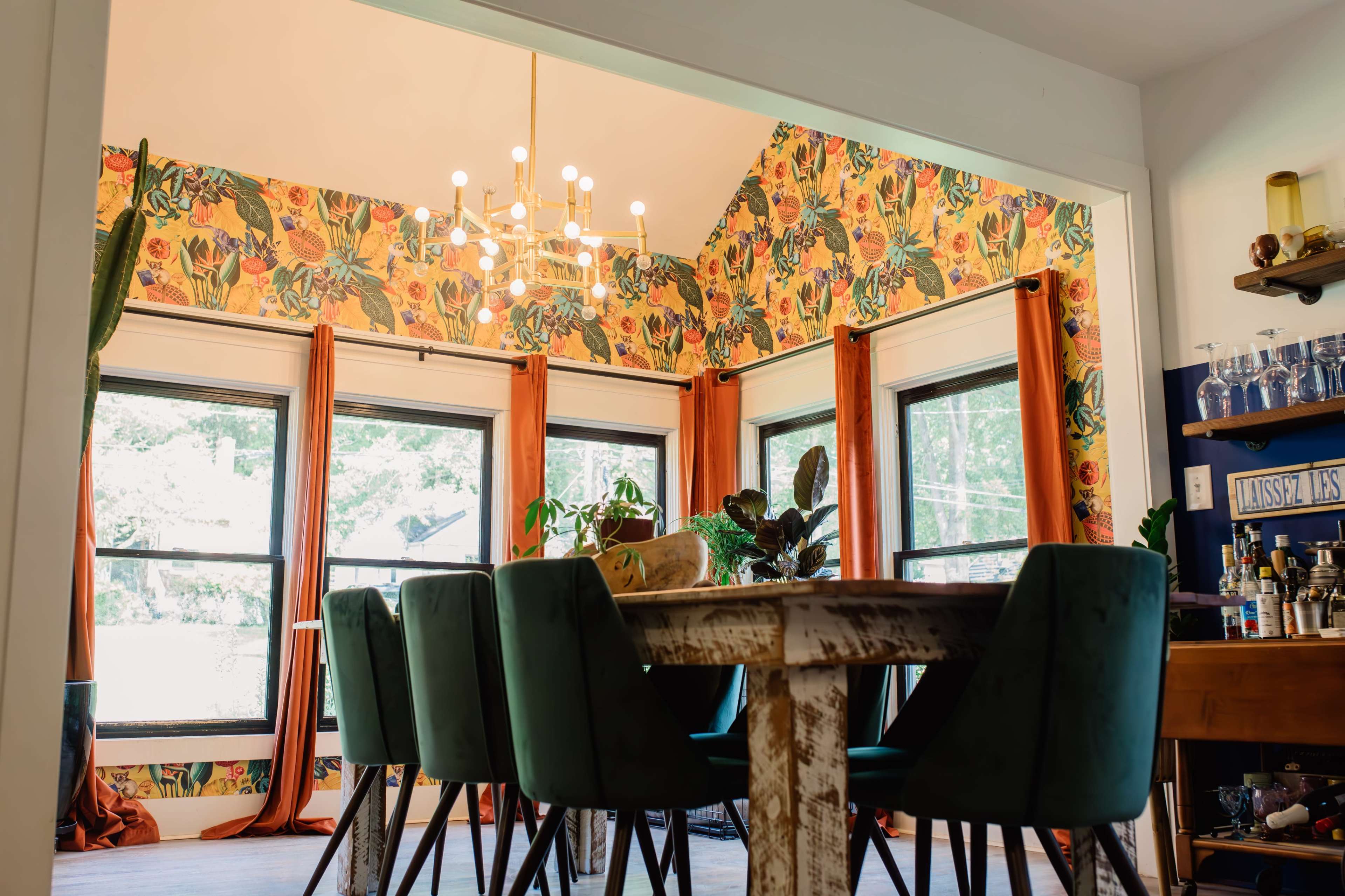 A dining area with a wooden table surrounded by green upholstered chairs, accented by floral wallpaper and large windows that provide natural light.