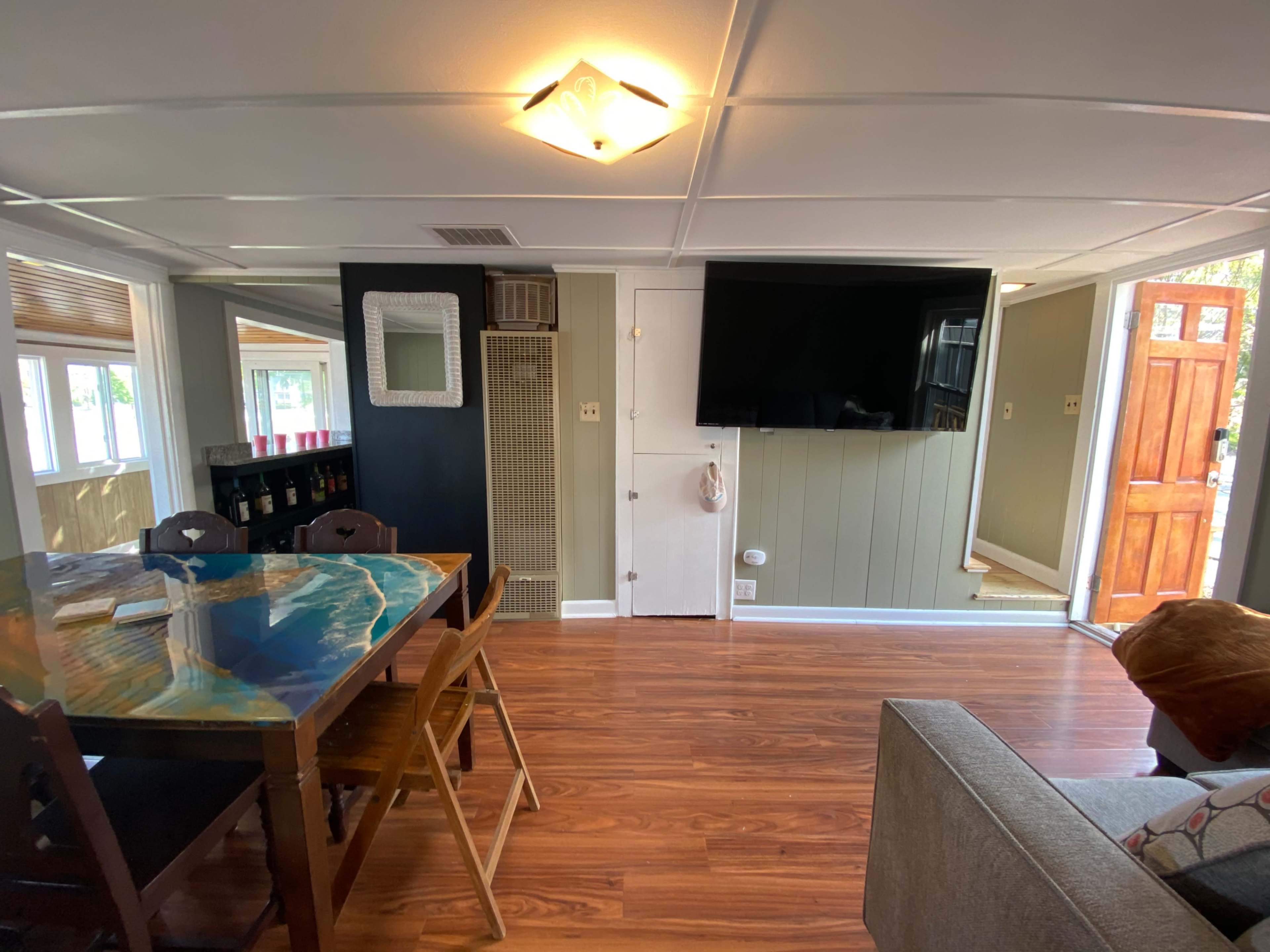 The scene shows a combined living and dining area featuring a table with a decorative top, a television mounted on the wall, and an entrance door leading outside.