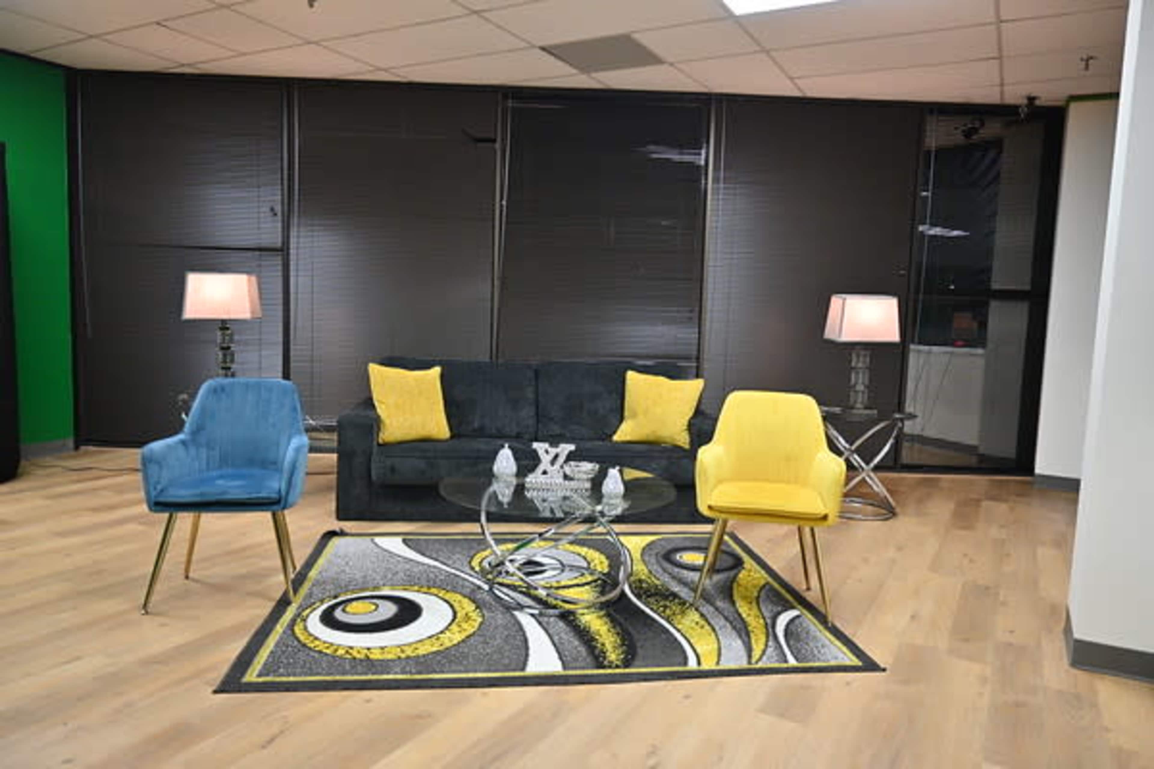 A modern living room setup featuring a black sofa, two colorful chairs, a round glass coffee table, and a patterned rug.