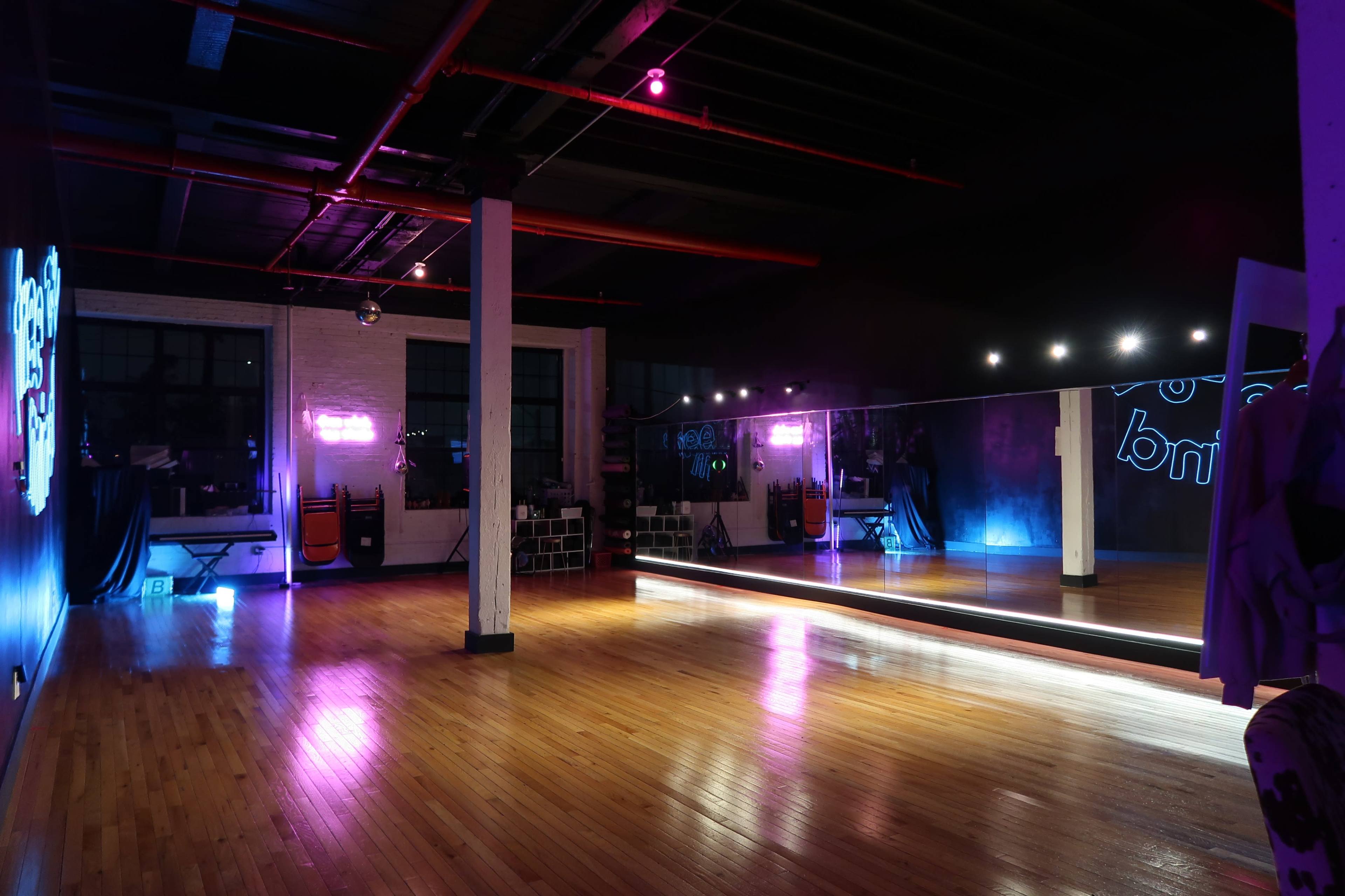 The image shows a spacious, dimly lit dance studio with polished wooden floors, a large mirror, and neon lights illuminating the walls.