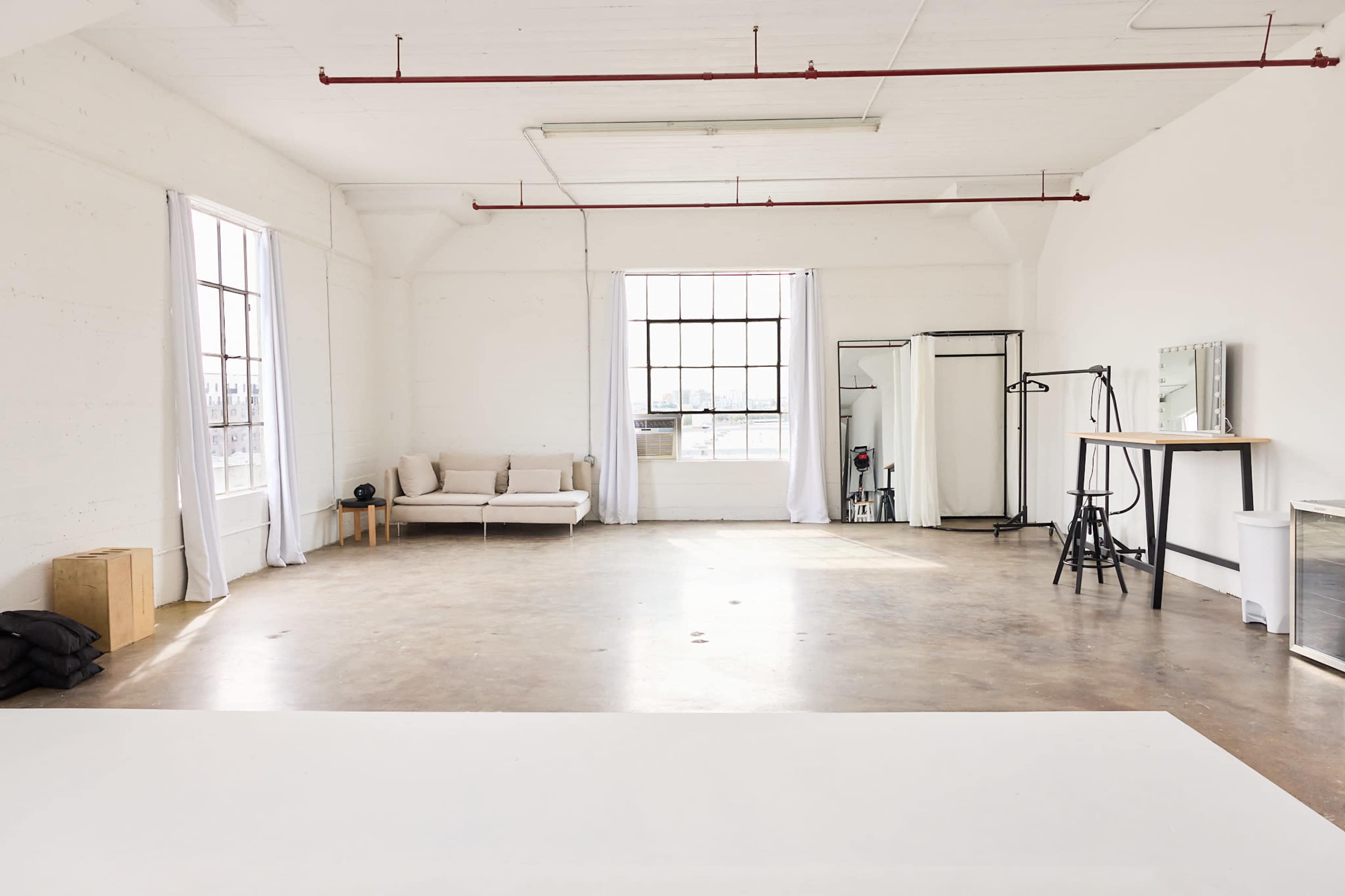 The image shows a spacious, minimalist studio with large windows, a couch, a mirror, and various furniture items arranged in a bright, open setting.