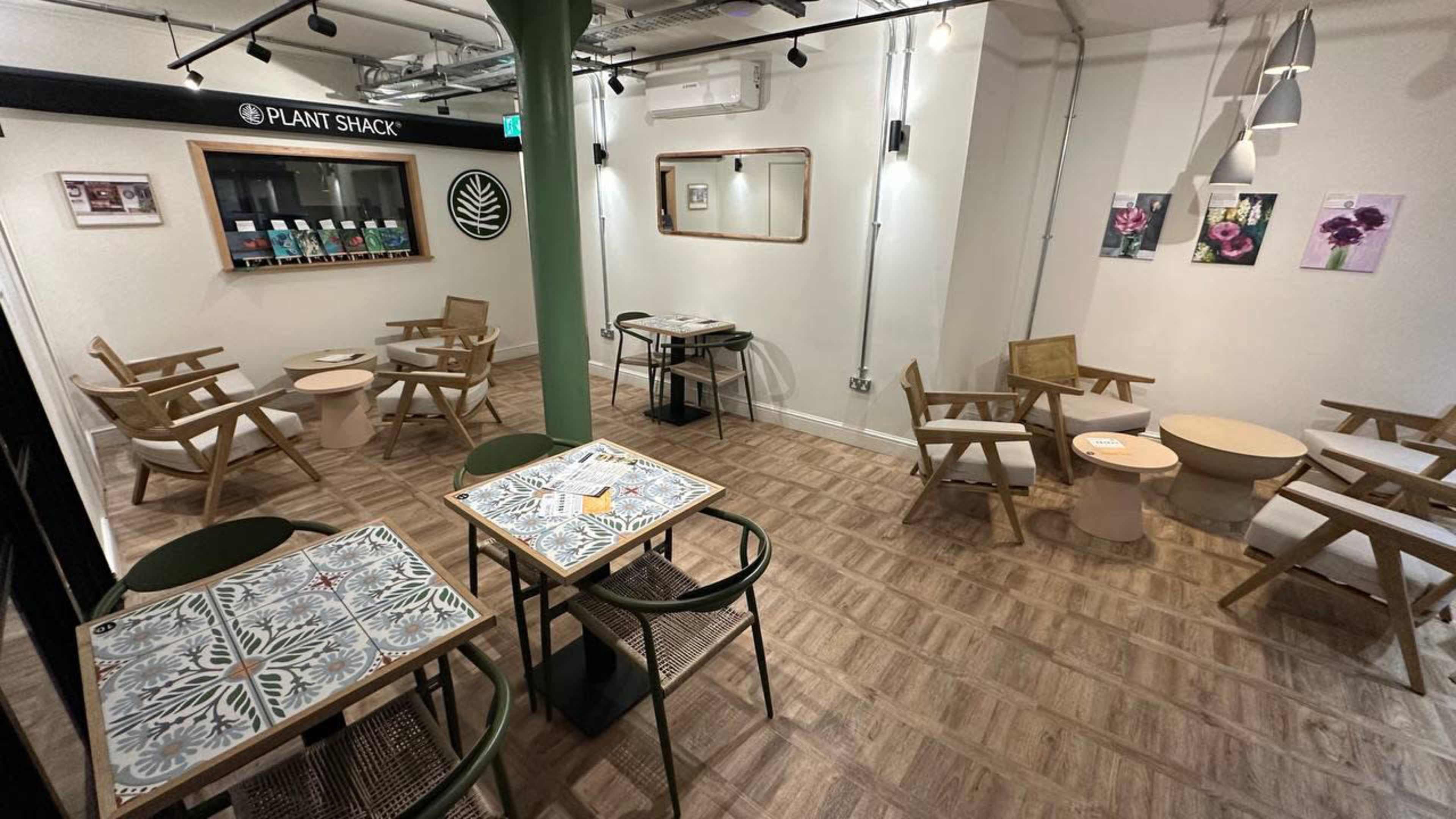 The image shows a modern café interior with several wooden tables and chairs arranged around a central area, featuring green accents and decorative wall art.