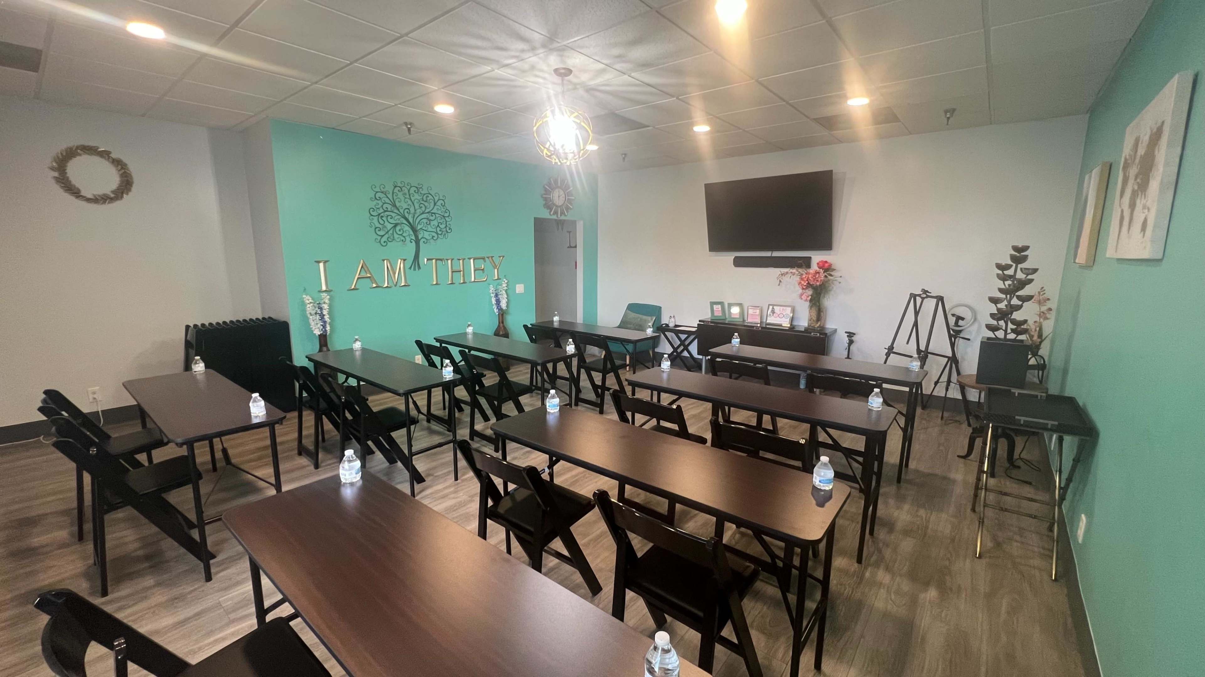A spacious meeting room arranged with rows of black folding tables and chairs, featuring a television on the front wall and decorations that include a tree design and the words "I AM THEY."