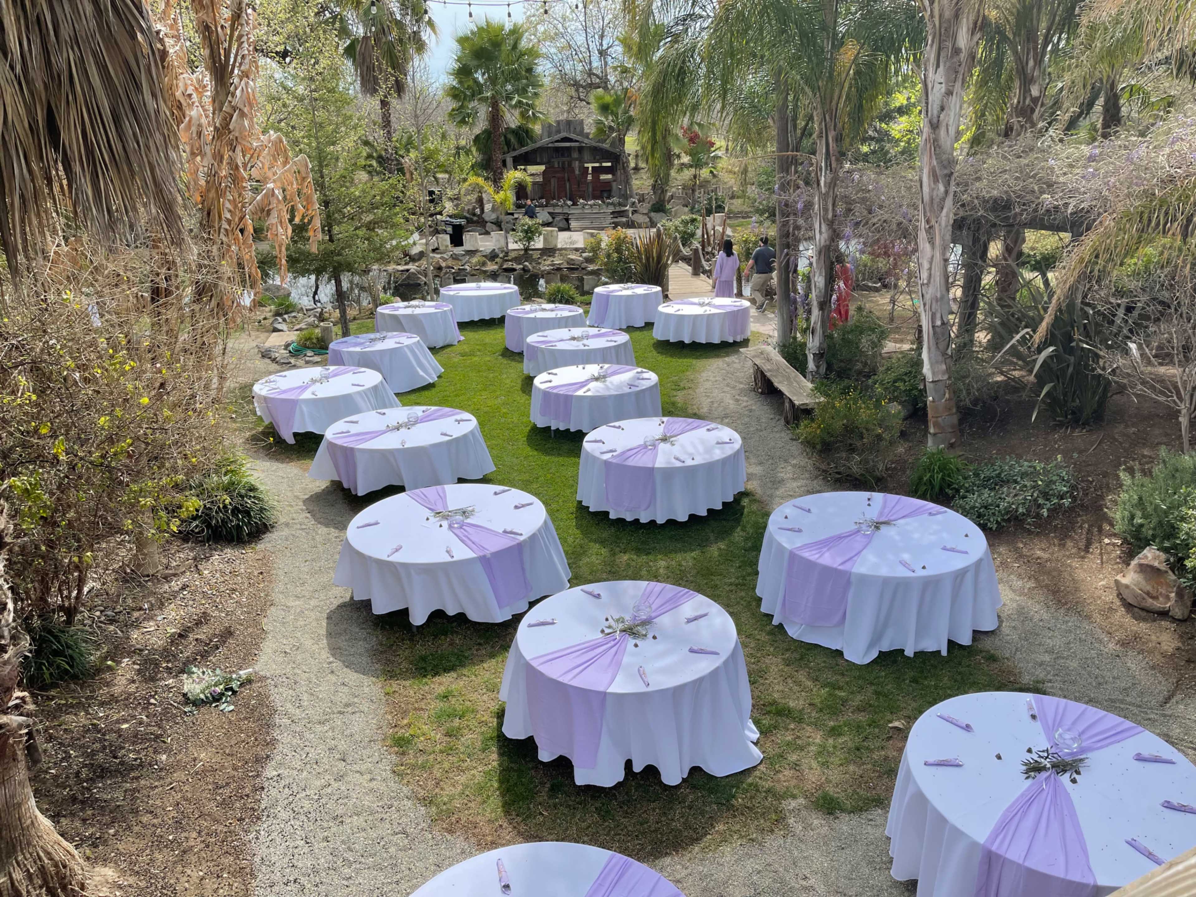 A garden venue set up for an outdoor event with round tables covered in white tablecloths and lavender accents arranged on a grassy area.