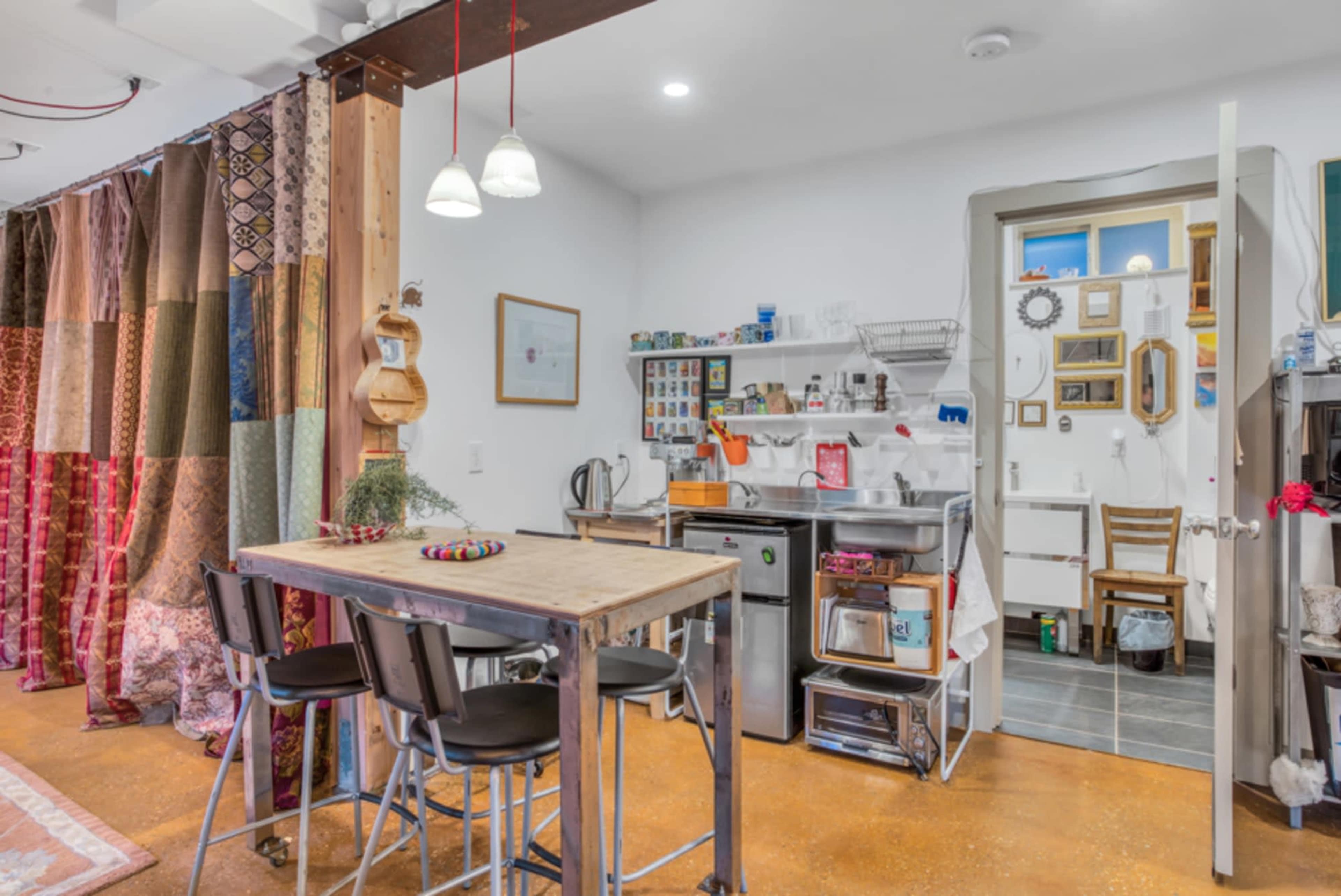 A small kitchen area with a wooden table, black chairs, and a variety of kitchen appliances and storage units against a backdrop of patterned curtains.