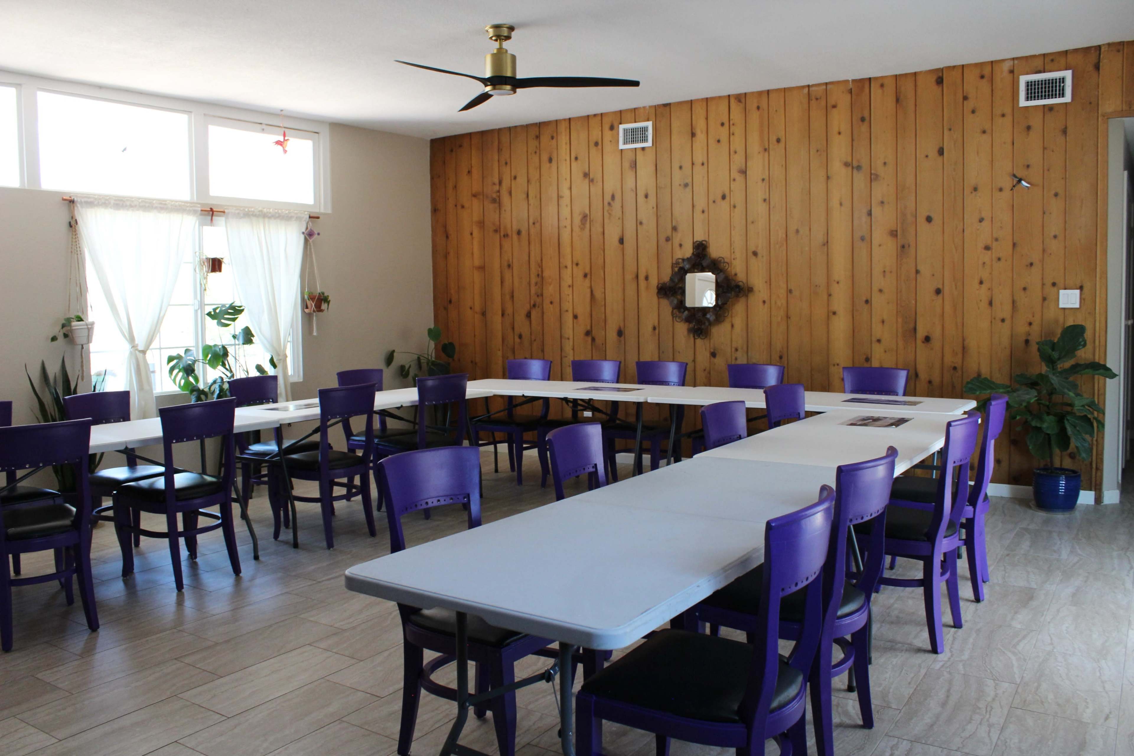 The image shows a spacious room with several tables arranged in a U-shape, surrounded by purple chairs and featuring wooden paneling on one wall.