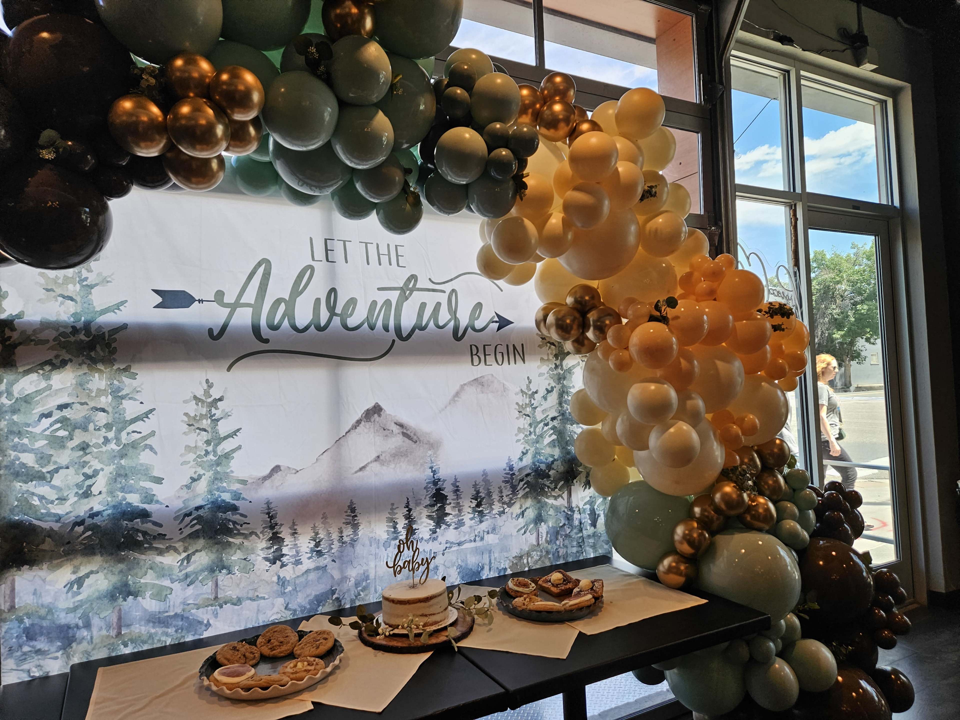 A decorative display featuring a balloon arch and a dessert table is set against a backdrop with a mountain scene and the words "Let the Adventure Begin."
