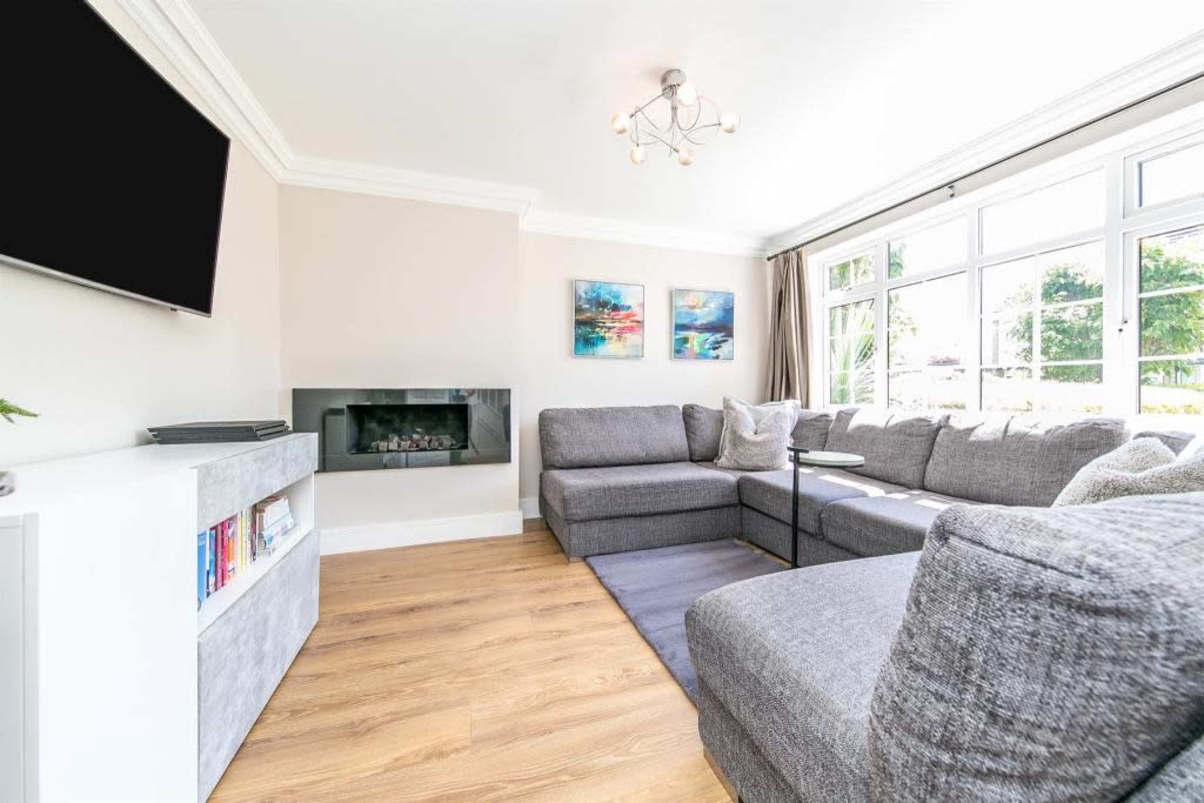 A modern living room with a sectional gray sofa, a wall-mounted television, a sleek electric fireplace, and large windows allowing natural light to enter.