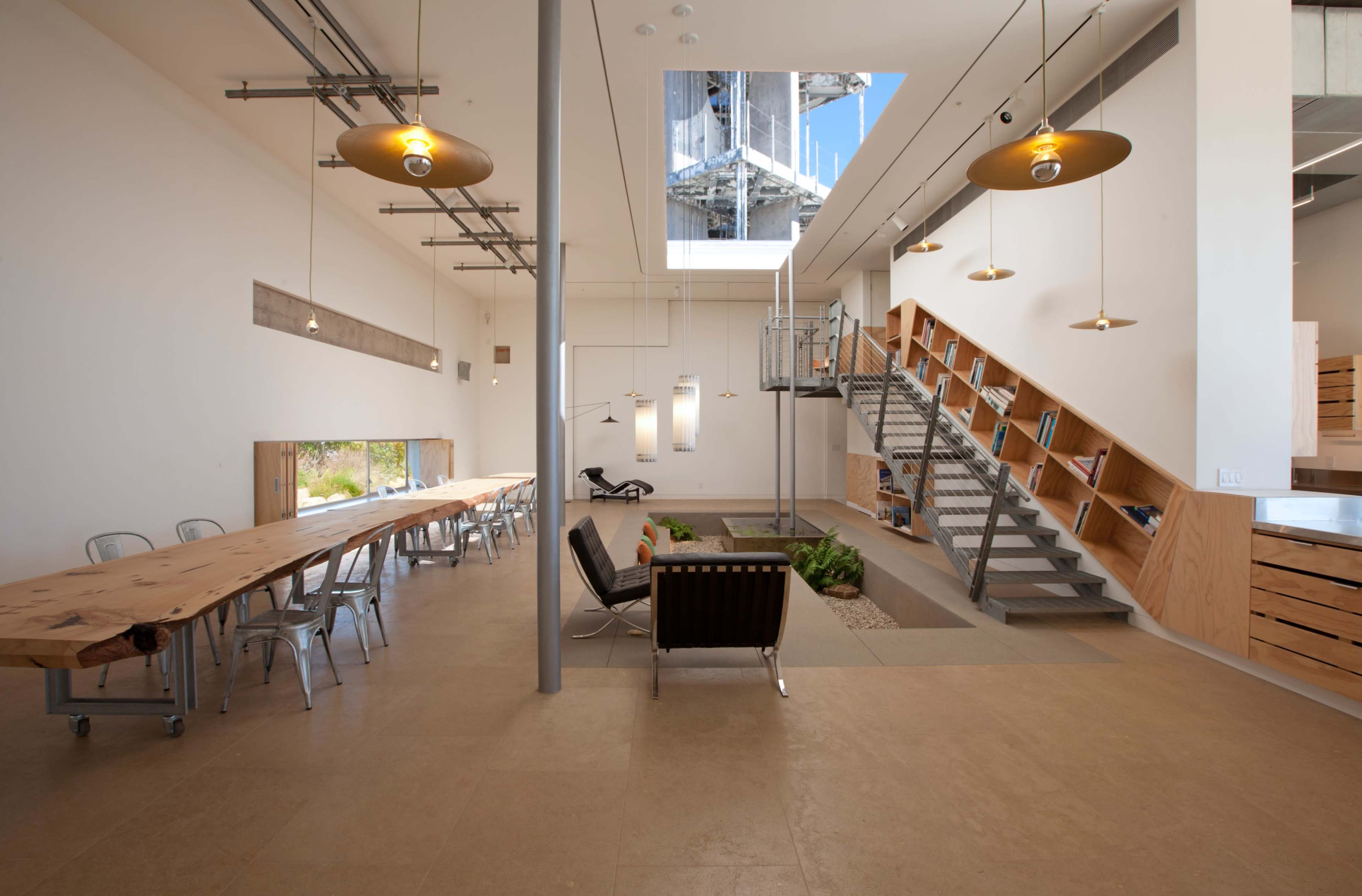 The image shows a modern, spacious interior of an office or workspace featuring a large wooden table, a staircase leading to an upper level, and a glass skylight illuminating the area.