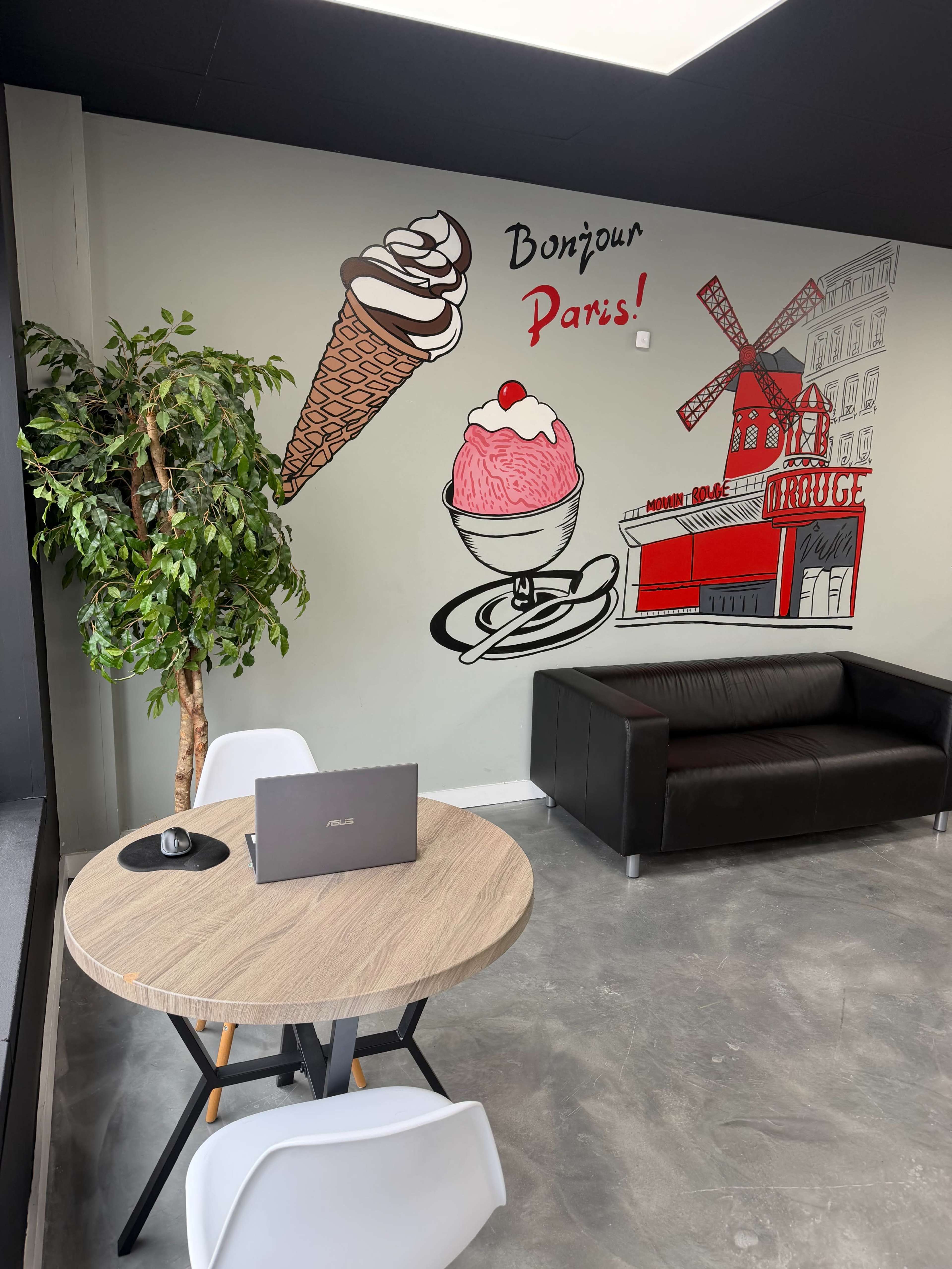 The image shows a modern interior space featuring a round wooden table with a laptop, a black leather sofa, and a wall decorated with illustrations of ice cream and a windmill, along with the text "Bonjour Paris!"