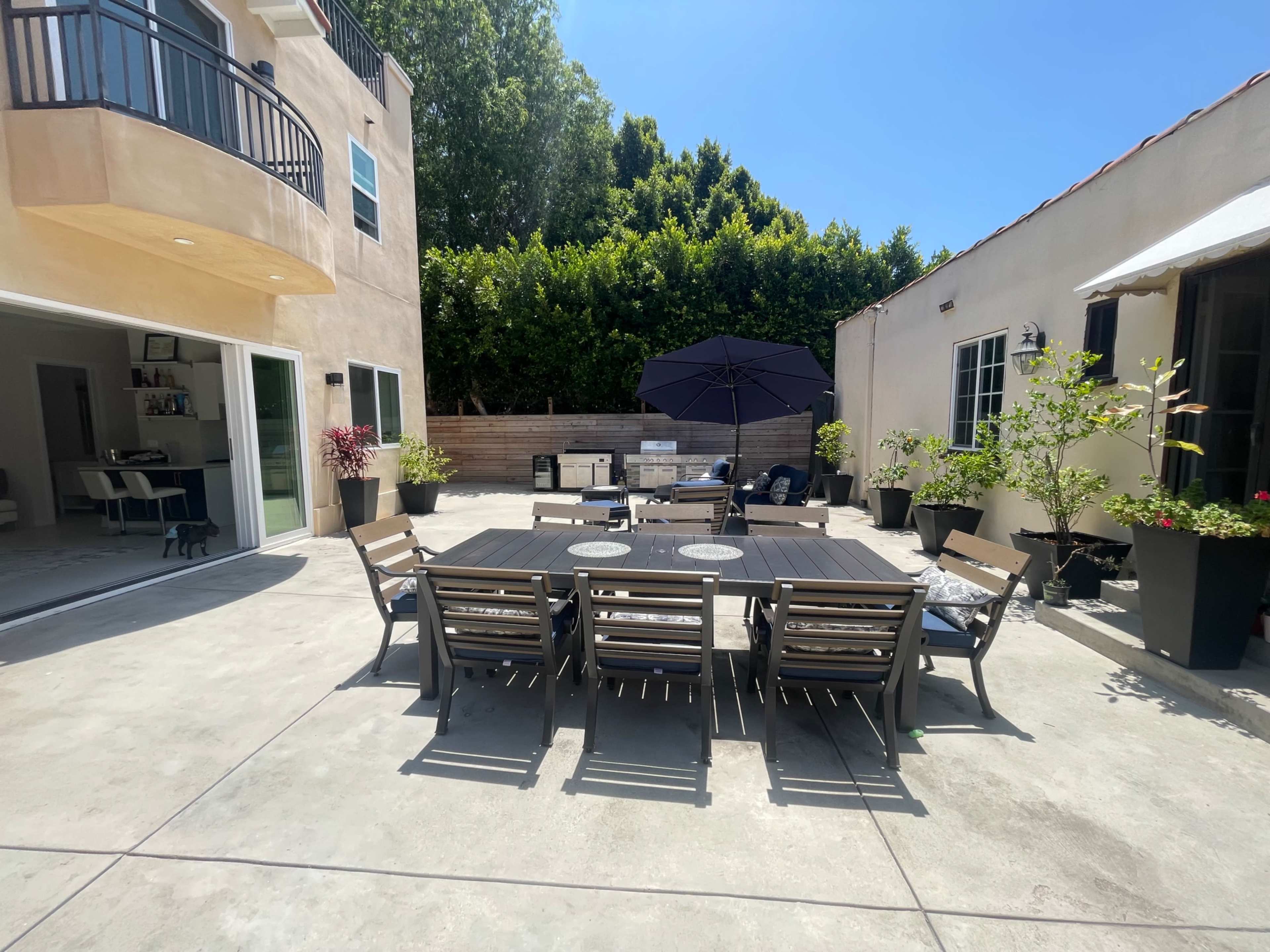 The image shows a spacious outdoor patio area with a dining table surrounded by chairs, an umbrella, and potted plants, adjacent to two buildings.