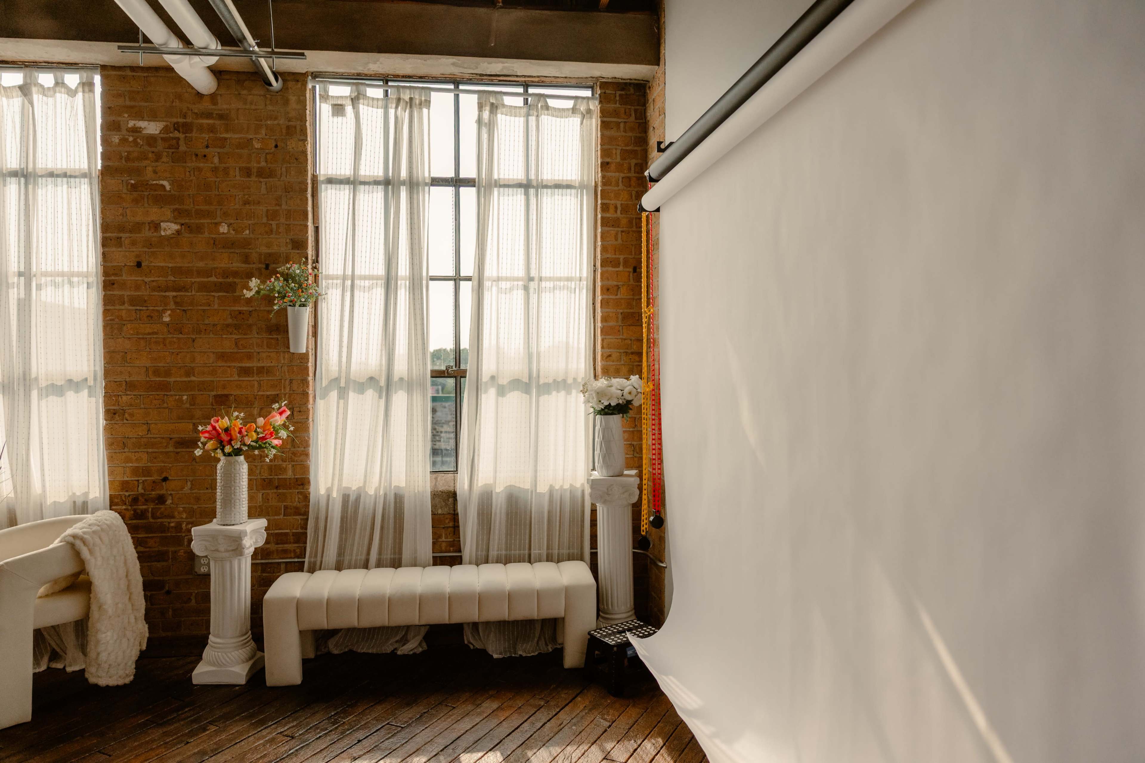 The image shows a bright room with brick walls, large windows covered by sheer curtains, and a white backdrop with decorative flower arrangements on pedestals.