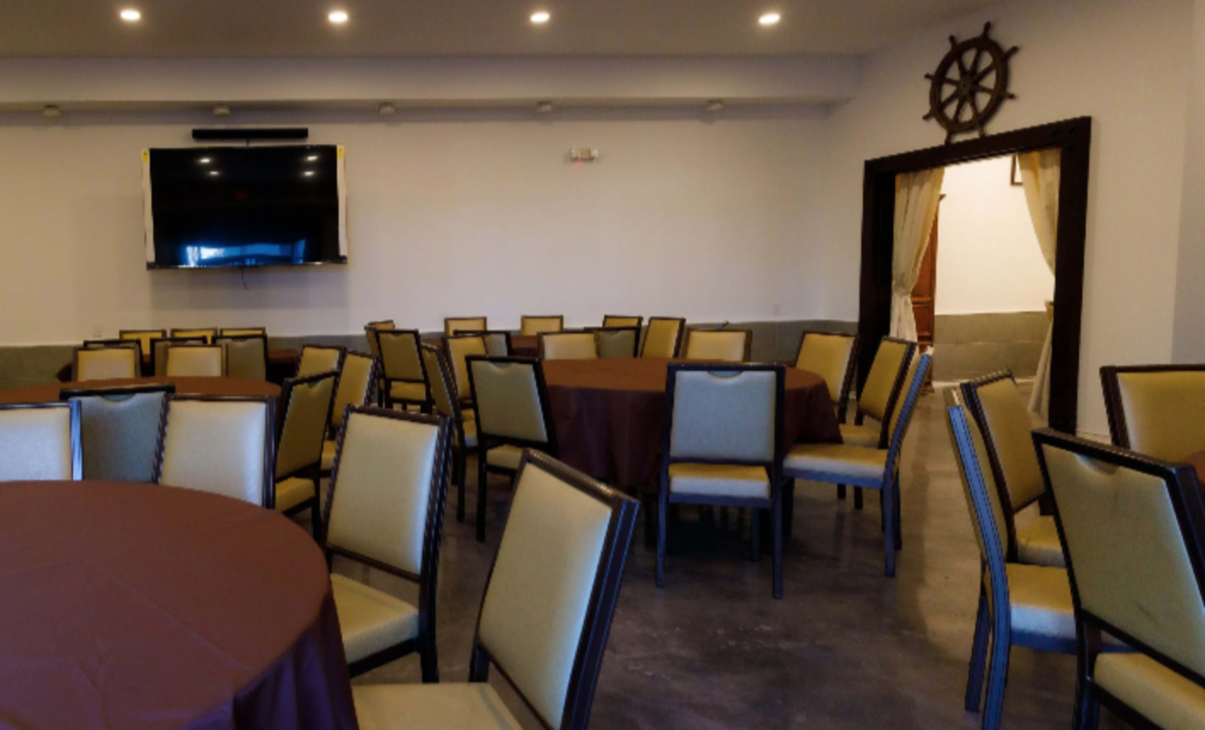 The image shows a spacious event room with multiple round tables covered in brown tablecloths and beige chairs arranged around them, alongside a television mounted on the wall.