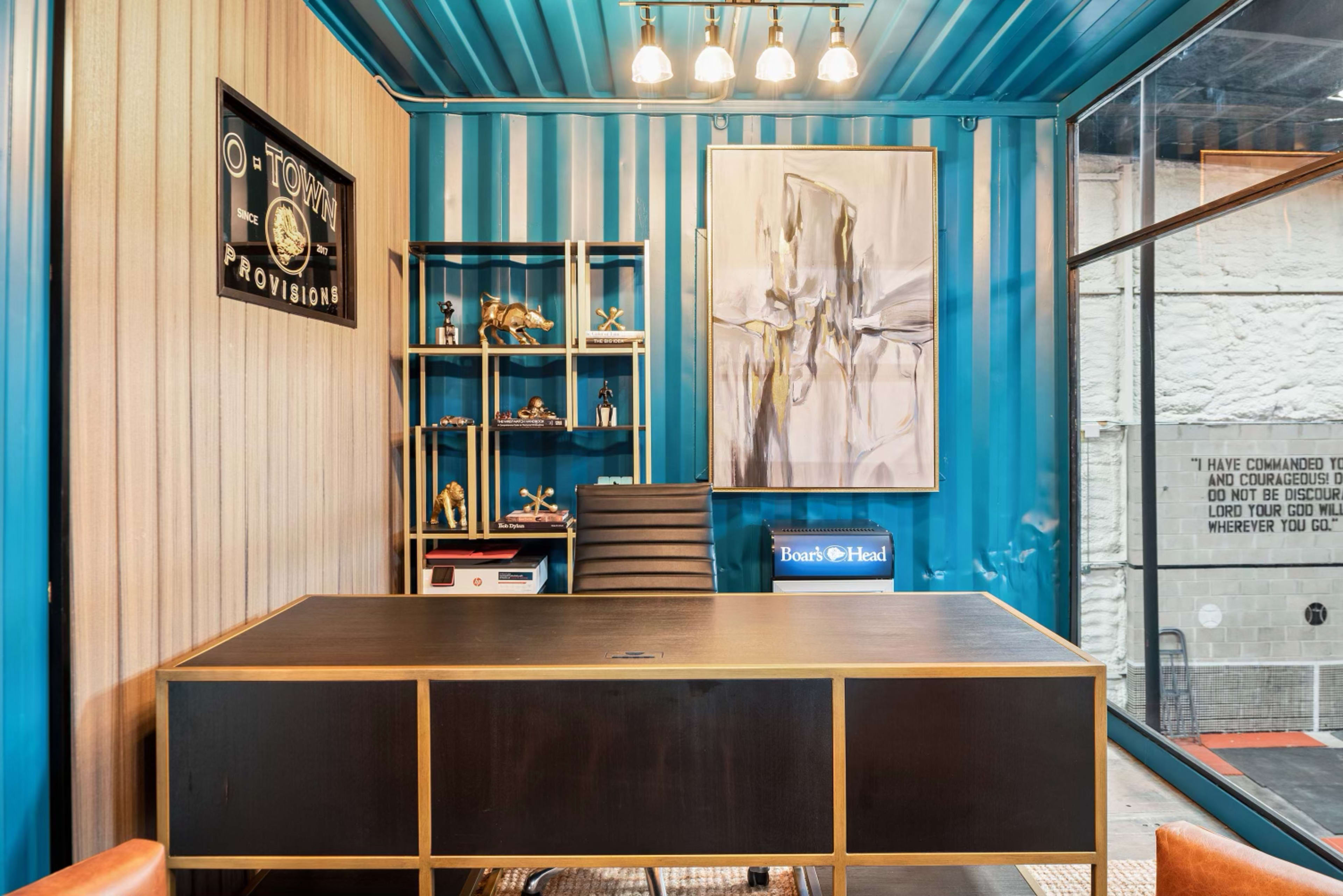 The image shows a modern office space featuring a large black desk, colorful blue and beige walls, a shelf with decorative items, and a piece of abstract art hanging on the wall.