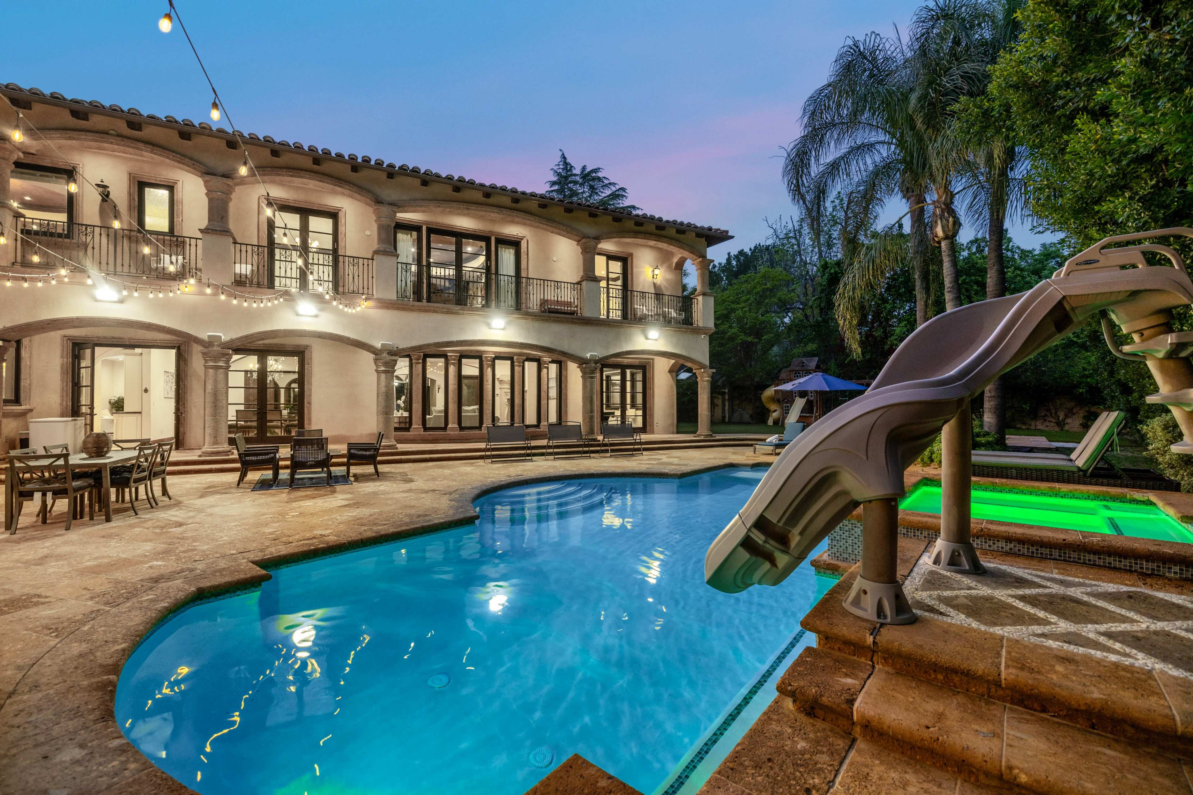 A spacious backyard with a swimming pool, a water slide, and a two-story house featuring multiple balconies and outdoor seating areas.
