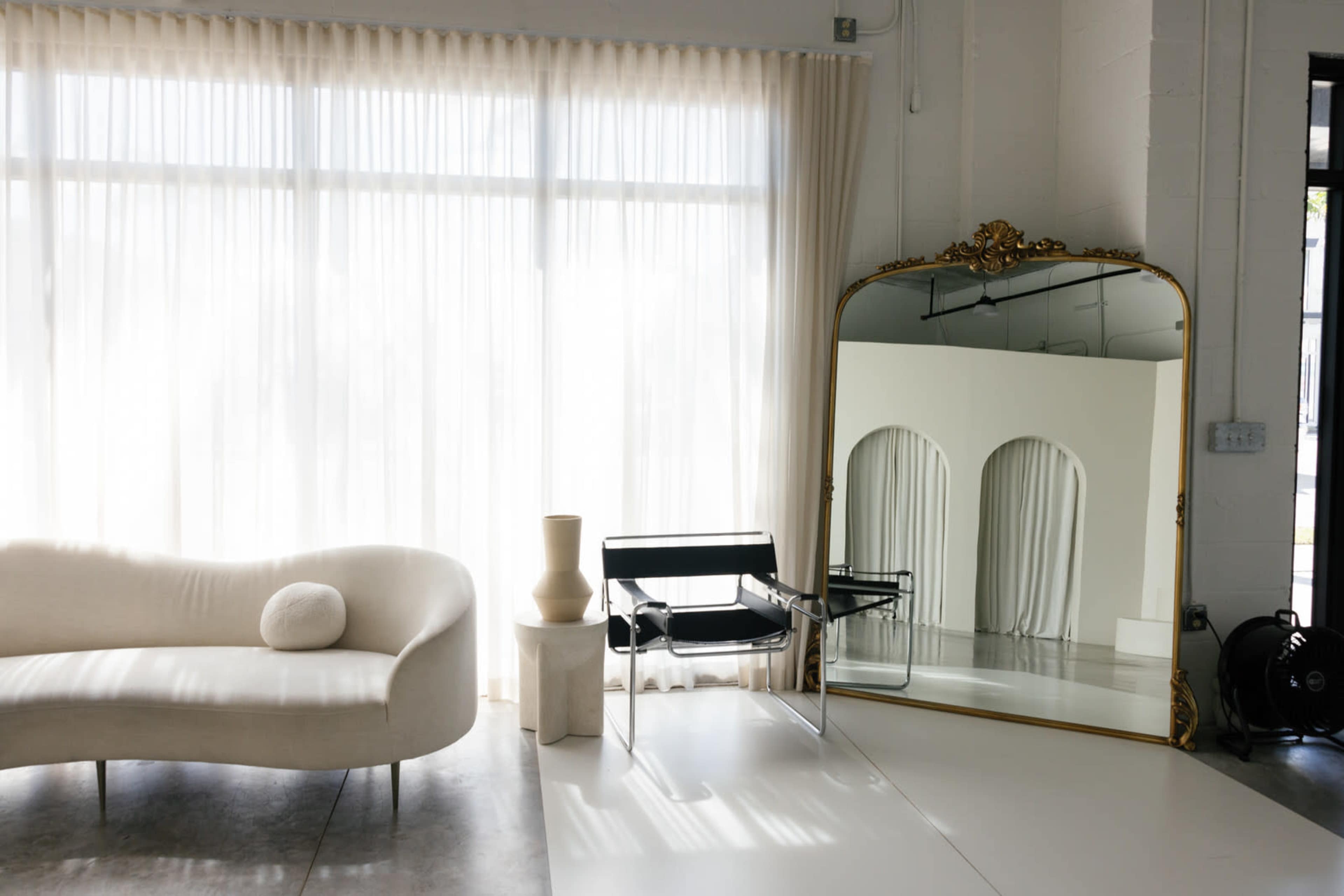 A modern interior features a curved sofa, a minimalist chair, a sculptural vase, and a large ornate mirror reflecting sheer curtains.