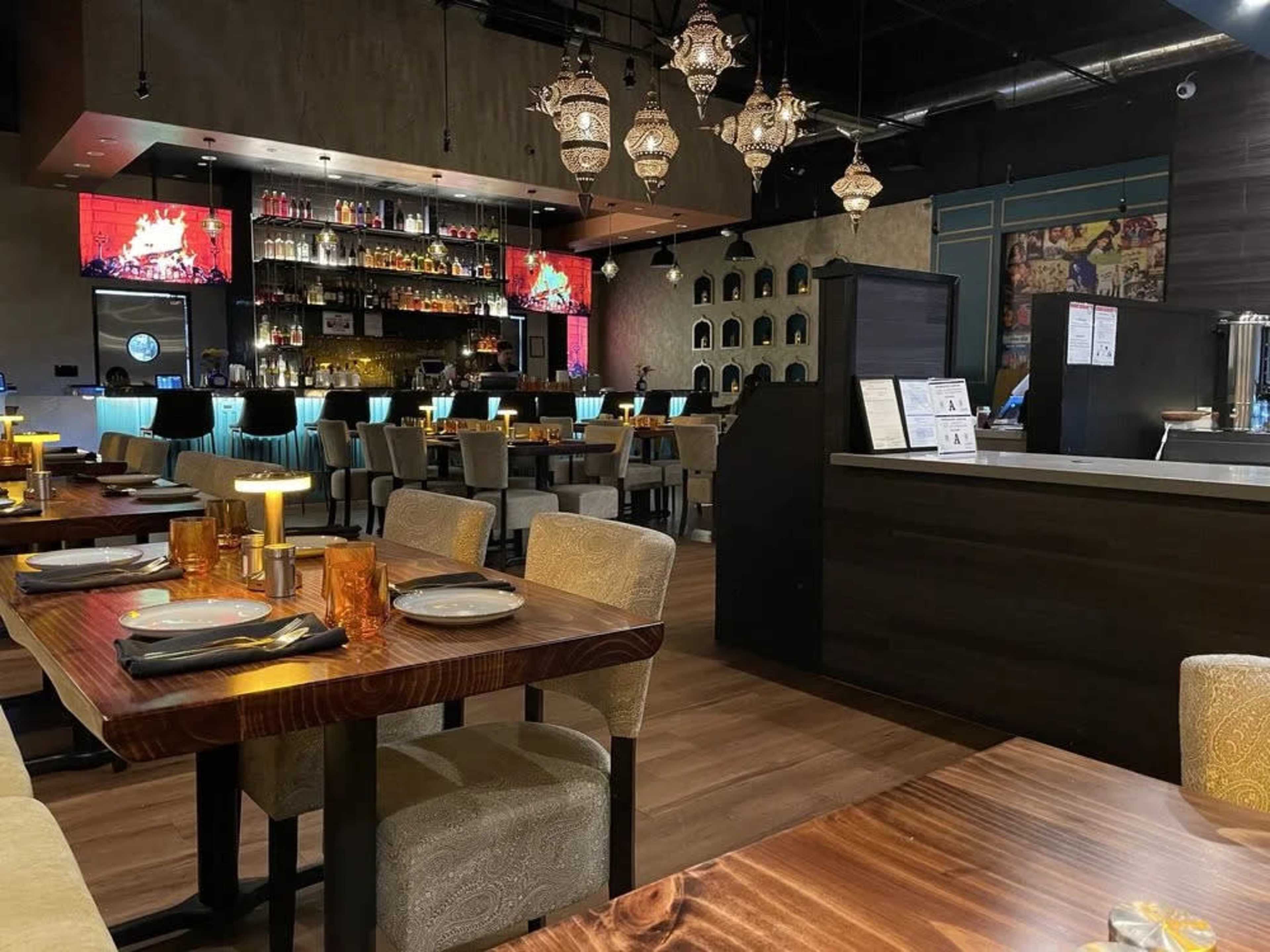 The image shows a modern restaurant interior with wooden tables, upholstered chairs, and a bar area featuring shelves of bottles and decorative lighting.