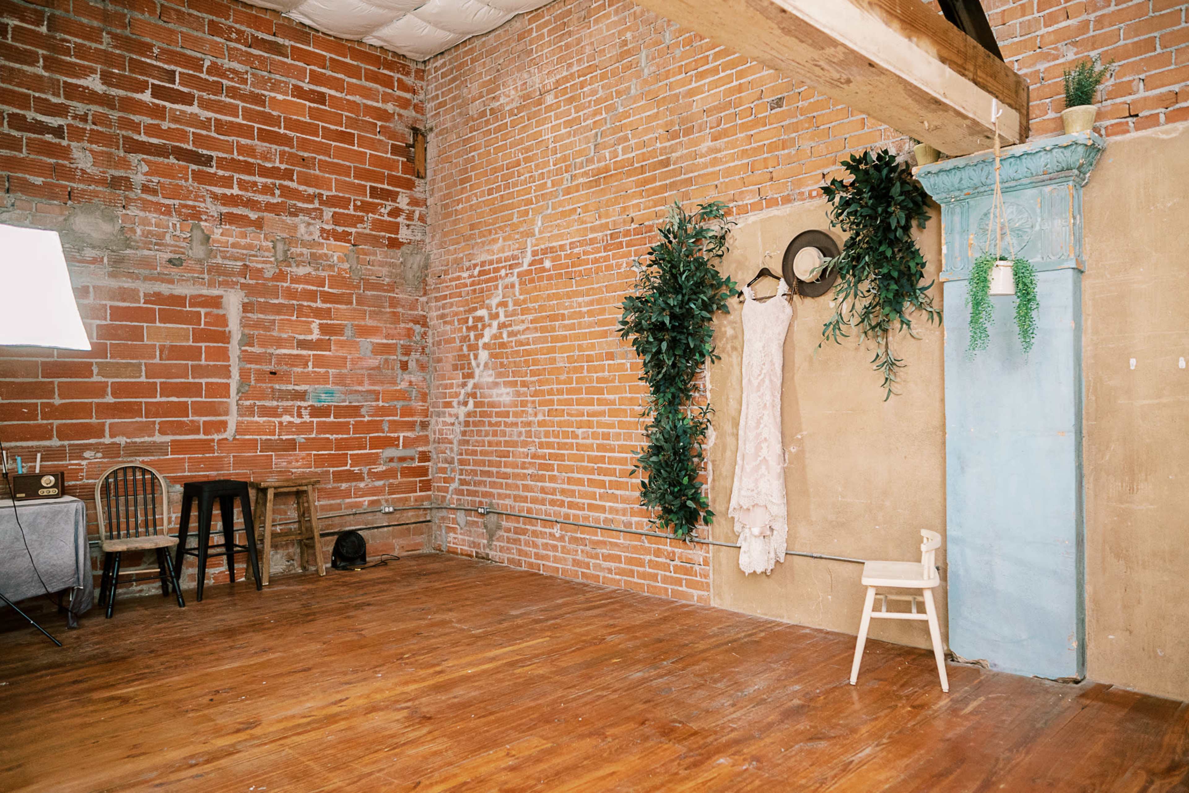 A spacious, unfurnished room with exposed brick walls, a wooden floor, a small table with chairs, and a hanging dress next to a blue decorative column.