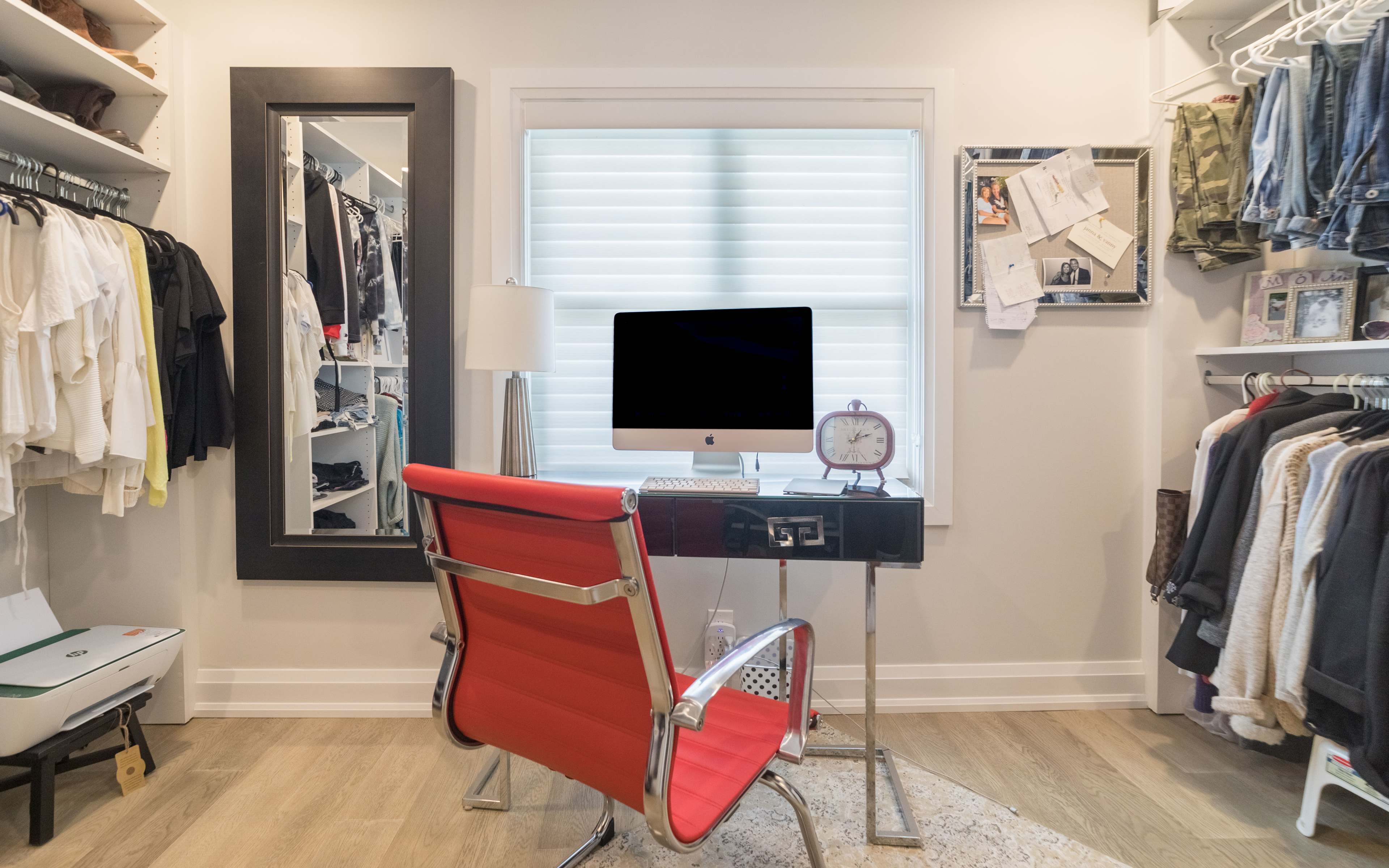 A modern home office is set up in a walk-in closet, featuring a desk with a computer, a red chair, and organized clothing on racks.