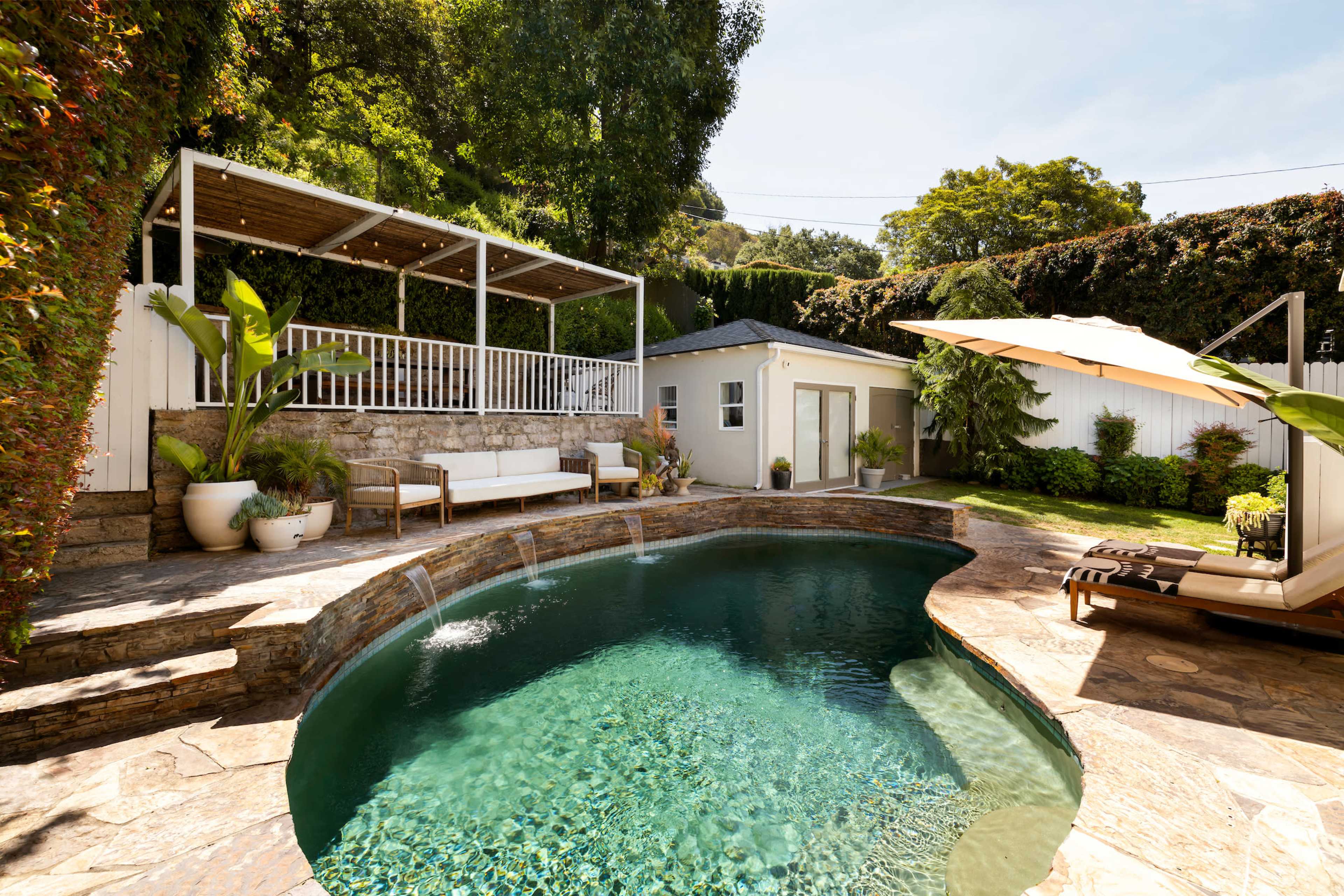 A modern, landscaped backyard features a small pool, stone seating areas, and a shaded lounge space surrounded by lush greenery.