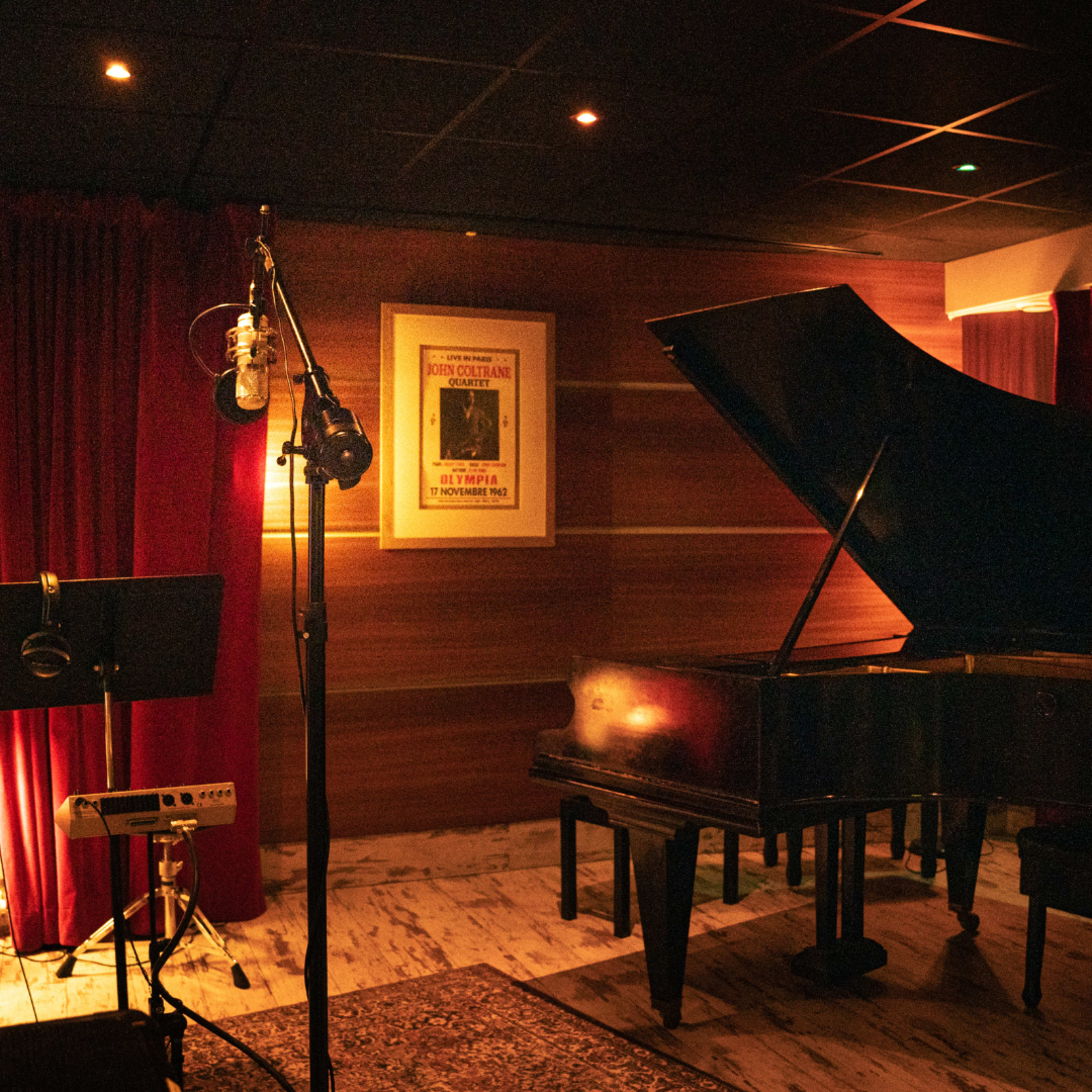 A grand piano sits beside a microphone and music stand in a dimly lit recording studio adorned with red curtains and framed artwork.