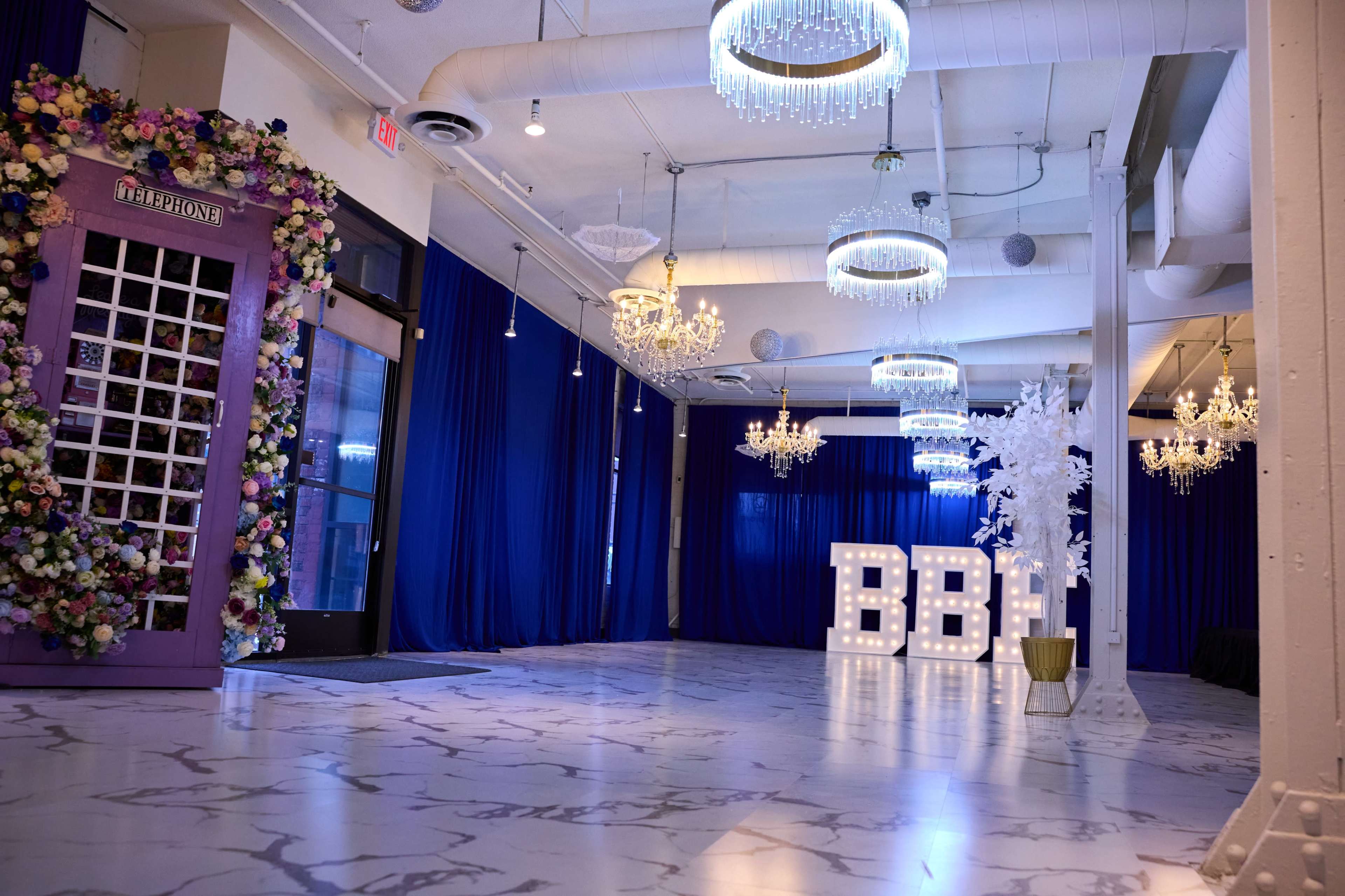 The image shows a decorated event space featuring purple and floral accents, elegant chandeliers, and large illuminated letters spelling "BBI" against a blue backdrop.