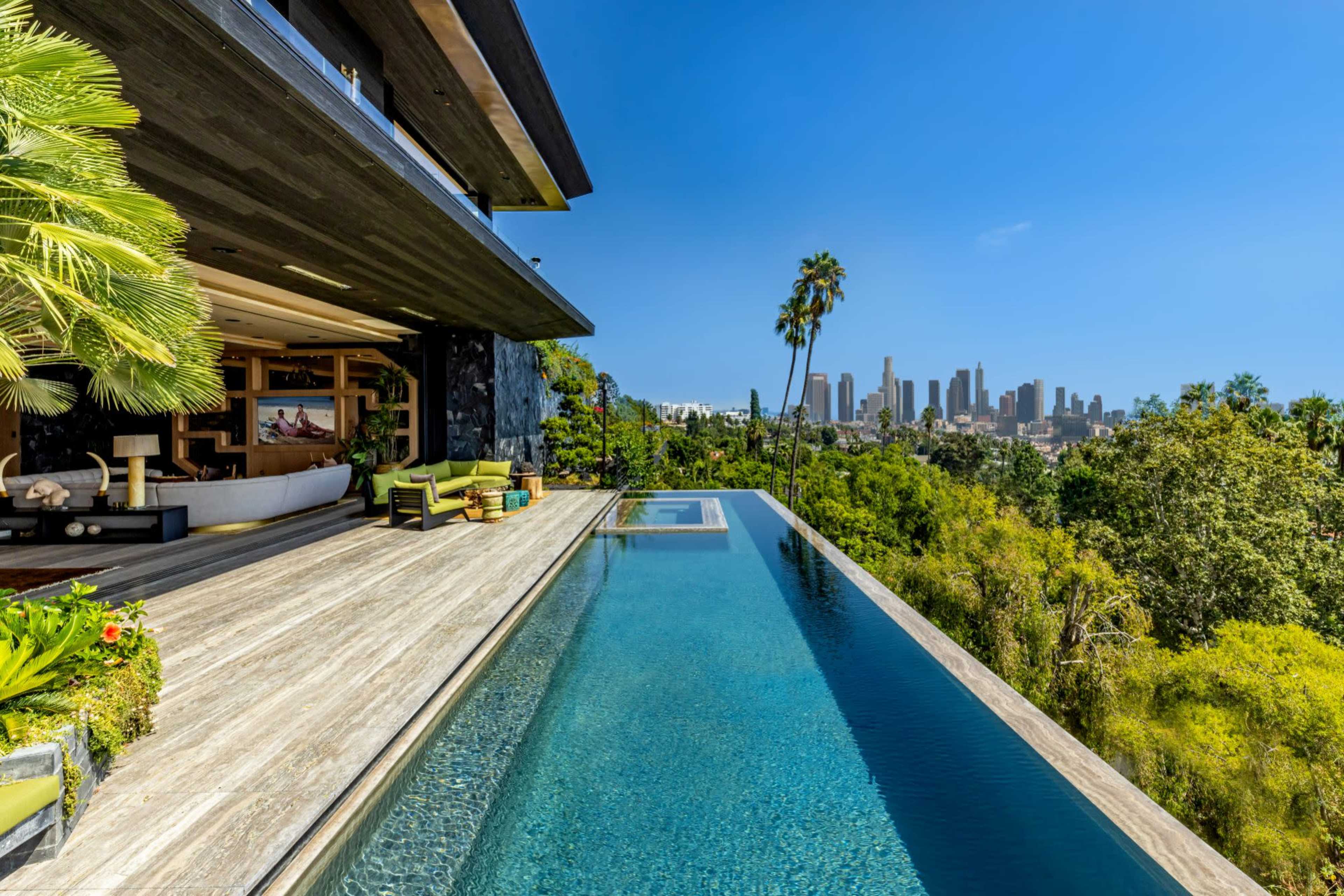 A luxurious modern home with a large infinity pool overlooks a city skyline surrounded by greenery.