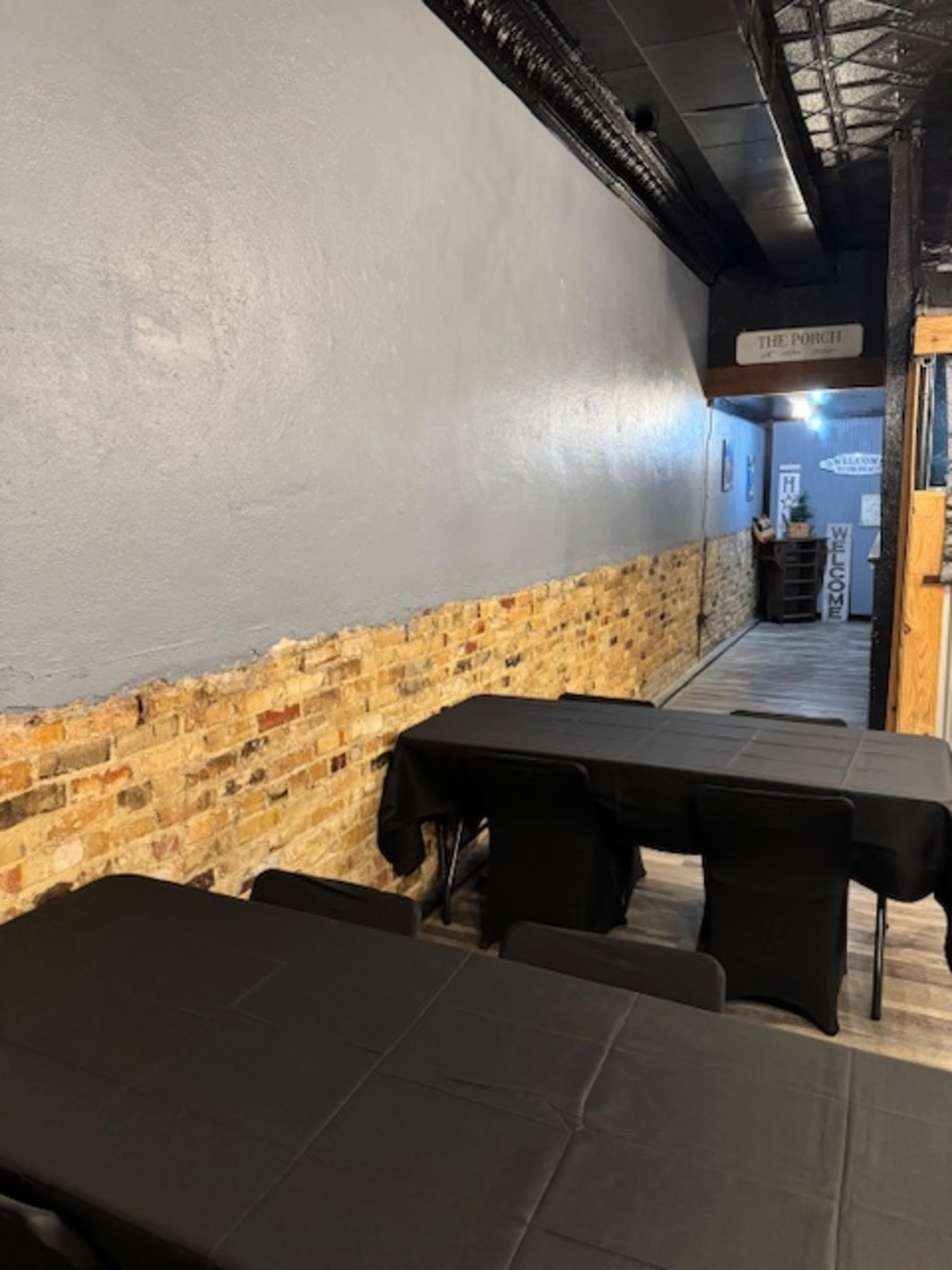 The image shows a narrow hallway with exposed brick walls and several black-covered tables set up along one side.